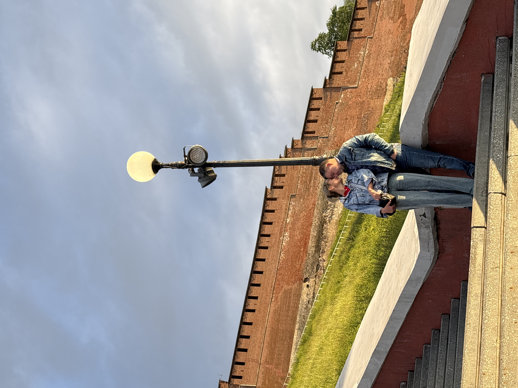 A couple is standing on steps, kissing, near a tall lamppost. They are both wearing denim jackets and jeans. Behind them is a tall brick wall with battlements and a grassy slope. The location is likely the Nizhny Novgorod Kremlin, a historic fortress in Nizhny Novgorod, Russia. The sky is cloudy with some patches of blue.