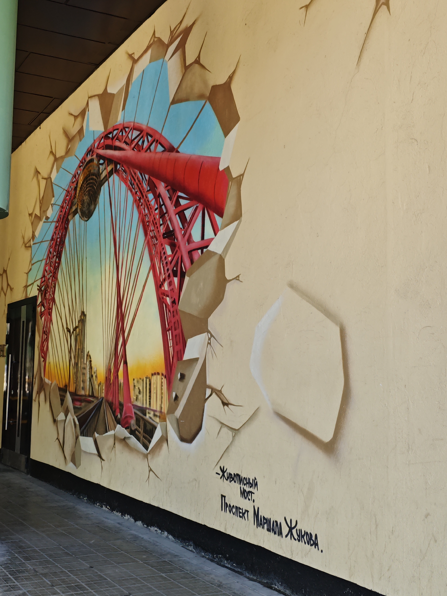 The image shows a mural painted on the exterior wall of a building in Moscow, Russia. The mural depicts a realistic, three-dimensional effect of a bridge breaking through the wall, with a section of the red suspension bridge and its cables visible against a sky with a gradient from light blue to orange. In the background, there is a cityscape with buildings. The mural is signed with Russian text: "- Живописный МОСТ. Проспект Маршала Жукова." This translates to "- Picturesque BRIDGE. Marshal Zhukov Avenue." The bridge depicted is likely the Trekhgory Bridge, also known as the "Picturesque Bridge," located on Marshal Zhukov Avenue in Moscow. The foreground shows a tiled walkway leading away from the wall. The wall itself is painted a light beige color, with cracks and shadows artfully rendered to enhance the illusion of depth.