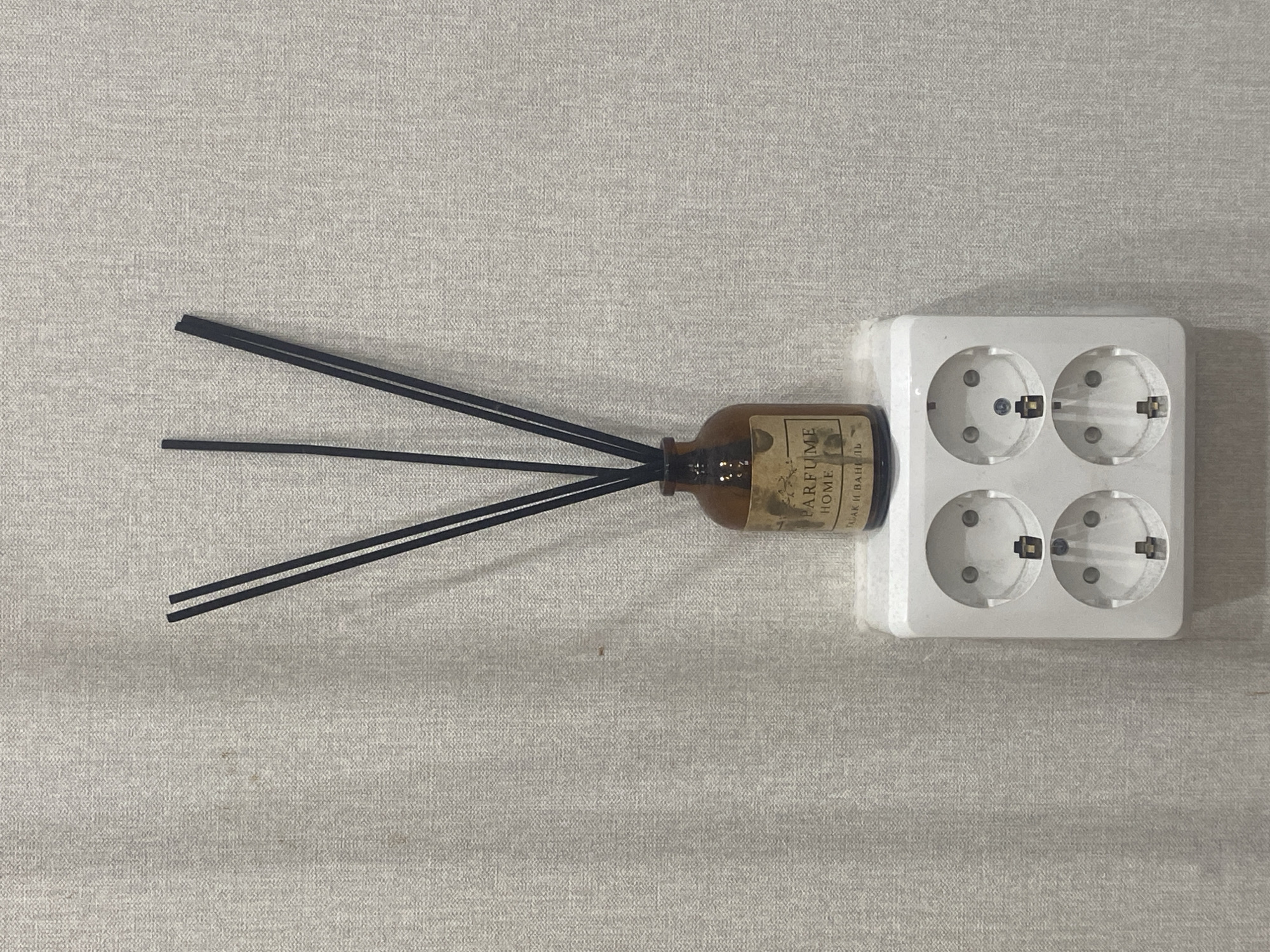 A brown glass diffuser bottle with black reeds stands on a white electrical outlet adapter. The bottle has a label that reads "PARFUME HOME" and some unreadable text below it. The outlet adapter is mounted on a textured, light-colored wall. The background is a plain, unadorned wall with a subtle woven pattern. There are no people or animals present. The scene appears to be an indoor domestic setting. The location in Dolgoprudny, Russia, is not determinable from the image itself.