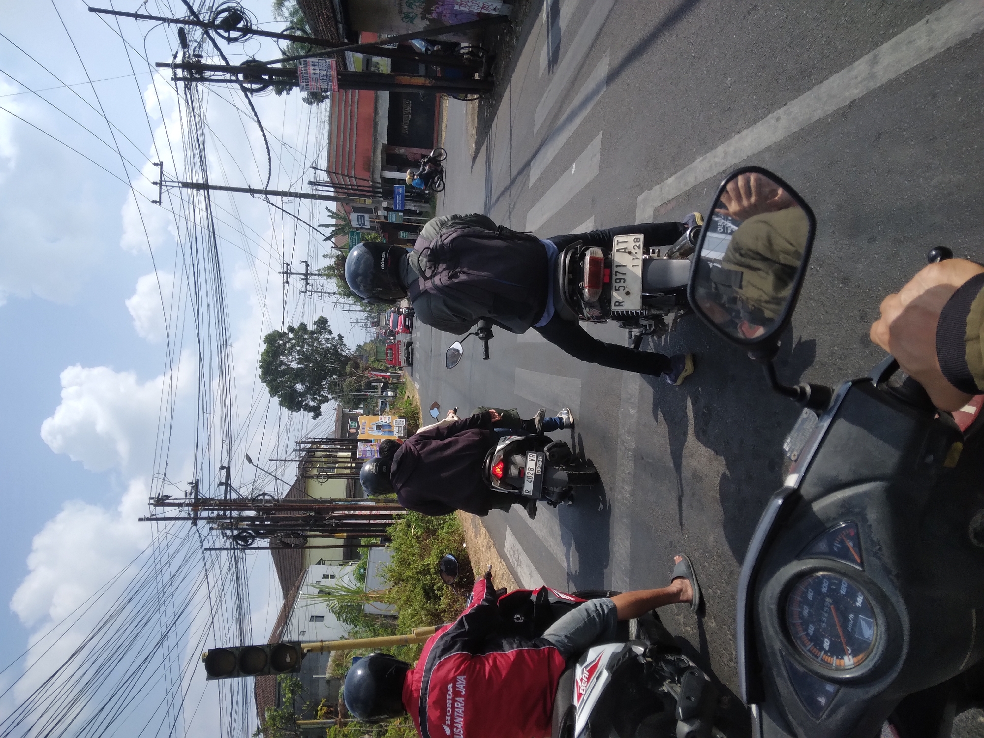The image captures a street scene in Mersi, Indonesia, likely during daylight hours under a partly cloudy sky. Several motorcycles are visible on the road, some with riders, indicating moderate traffic. In the foreground, the dashboard and handlebars of a motorcycle are prominent, suggesting the image was taken from the perspective of a rider. The road appears to be asphalt, with pedestrian crossings marked.
Several individuals are riding motorcycles. The closest motorcycle in the foreground has a rider wearing a red and black jacket with "HONDA NUSANTARA JAYA" branding. Another motorcycle further down the road has a rider wearing a black hoodie and a backpack, with a passenger behind them. A third motorcycle has a rider in a dark jacket with a backpack, also with a passenger.
The environment includes utility poles with numerous electrical wires crisscrossing overhead, a common sight in many Indonesian urban or semi-urban areas. Buildings with red and white facades line the street, some appearing to be shops or residences. Greenery, including trees and bushes, is also present. Traffic lights and road signs are visible, contributing to the urban streetscape. The overall impression is a typical street scene in a busy Indonesian town or city.
