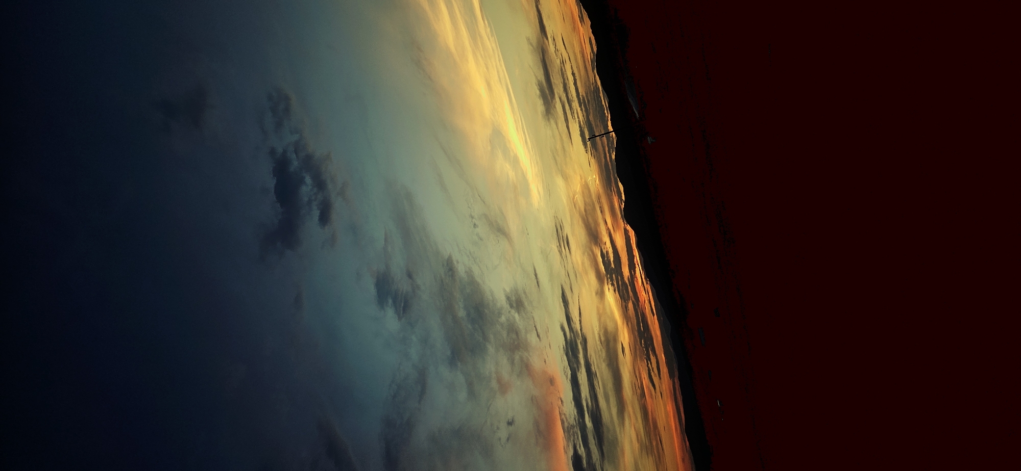 This is a vertical, slightly tilted photograph of a sky with a dramatic sunset over a dark landscape. The upper portion of the image is a deep, dark blue transitioning into teal and then a bright yellow and orange band where the sun is setting. Wispy clouds are visible throughout the sky, catching the light in various shades. Below the sky is a dark, silhouetted mountain range. In the foreground, the ground is a deep red or brown, also appearing silhouetted and indistinct. A thin, vertical dark object, possibly a stick or pole, is present in the middle ground, silhouetted against the sky. The image appears to be taken from an elevated position looking down towards the horizon. The location is identified as Momin Prohod, Bulgaria, suggesting a natural landscape setting, likely mountainous or hilly. The lighting indicates dusk or twilight.