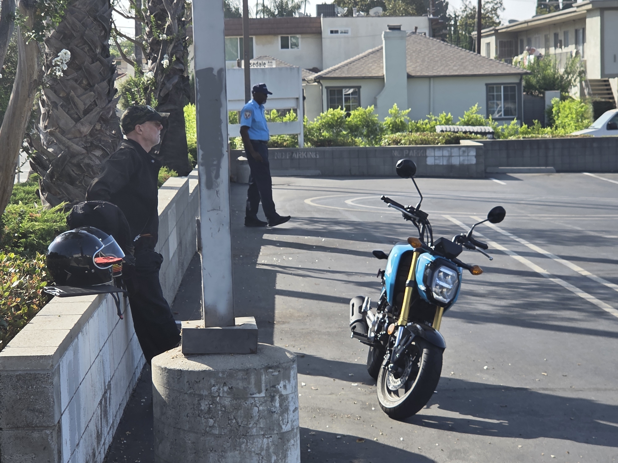 In a paved outdoor area, likely a parking lot, a man wearing a camouflage baseball cap and a black jacket stands near a textured concrete wall and a palm tree. He is looking to his right, with his left hand on his hip. A black motorcycle helmet rests on the wall in front of him.

In the background, another person, dressed in a light blue shirt and dark pants, stands facing away from the camera. This person appears to be wearing a hat and dark shoes. Behind them, a row of buildings with light-colored siding and dark roofs suggests a residential or commercial area. Green bushes and trees are visible around the buildings. The ground is asphalt, with distinct shadows indicating bright sunlight.

A bright blue and black motorcycle is parked in the foreground, facing towards the right of the frame. Its front wheel, handlebars, and headlight are clearly visible. The motorcycle has yellow front suspension forks.

The scene appears to be taking place during the daytime, under clear and sunny weather conditions. The presence of buildings and a paved area suggests an urban or suburban environment. There are no overt signs of a specific event, but the individuals' stances and the parked motorcycle could imply a brief stop or break. The overall mood seems relaxed and ordinary.

There is visible text on the gray concrete wall in the background which reads "NO PARKING". Another smaller sign in the distance has faded text that is difficult to read but may include street names.