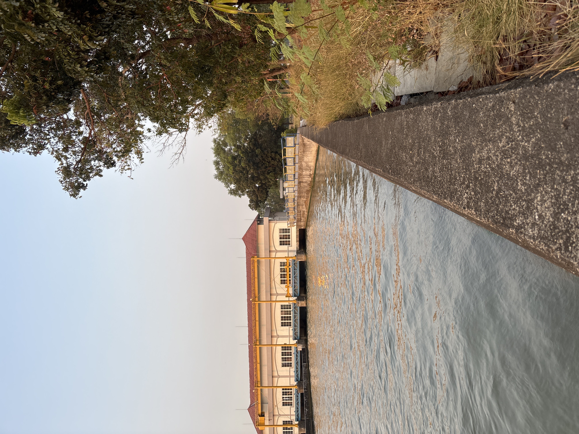 This image captures a waterfront view of a dam and its associated structures in Panyingkiran, Indonesia. The dam building, painted a light yellow with a red tiled roof, features numerous windows and appears to have mechanisms for controlling water flow, indicated by yellow cranes and blue gate structures. Adjacent to the dam, a concrete walkway runs along the water's edge, bordered by dry grass and vegetation on the right side of the frame. The water itself is calm, reflecting the warm tones of the sky, which transitions from a pale blue at the top to a lighter hue near the horizon. Lush green trees are visible in the background, some overhanging the walkway. The scene is devoid of any people, suggesting a tranquil, possibly early morning or late afternoon setting. The overall impression is one of an industrial water management facility integrated into a natural, semi-tropical environment.