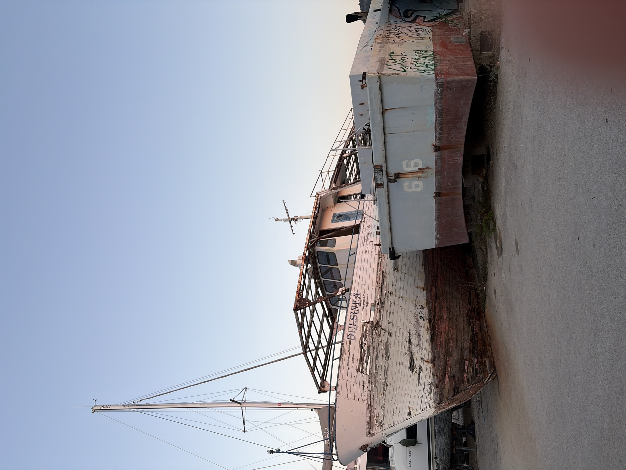 In the city of Bar, Montenegro, a wooden boat named "DULINEA" sits grounded on a dry dock. The boat is visibly weathered, with peeling white paint and exposed red wood. A metal frame structure is attached to the upper deck of the boat, showing signs of rust. To the right of the "DULINEA," a larger, lighter-colored vessel with graffiti on its side is also grounded. The sky above is clear and a pale blue, indicating it's likely early morning or late afternoon. The foreground consists of a concrete surface.