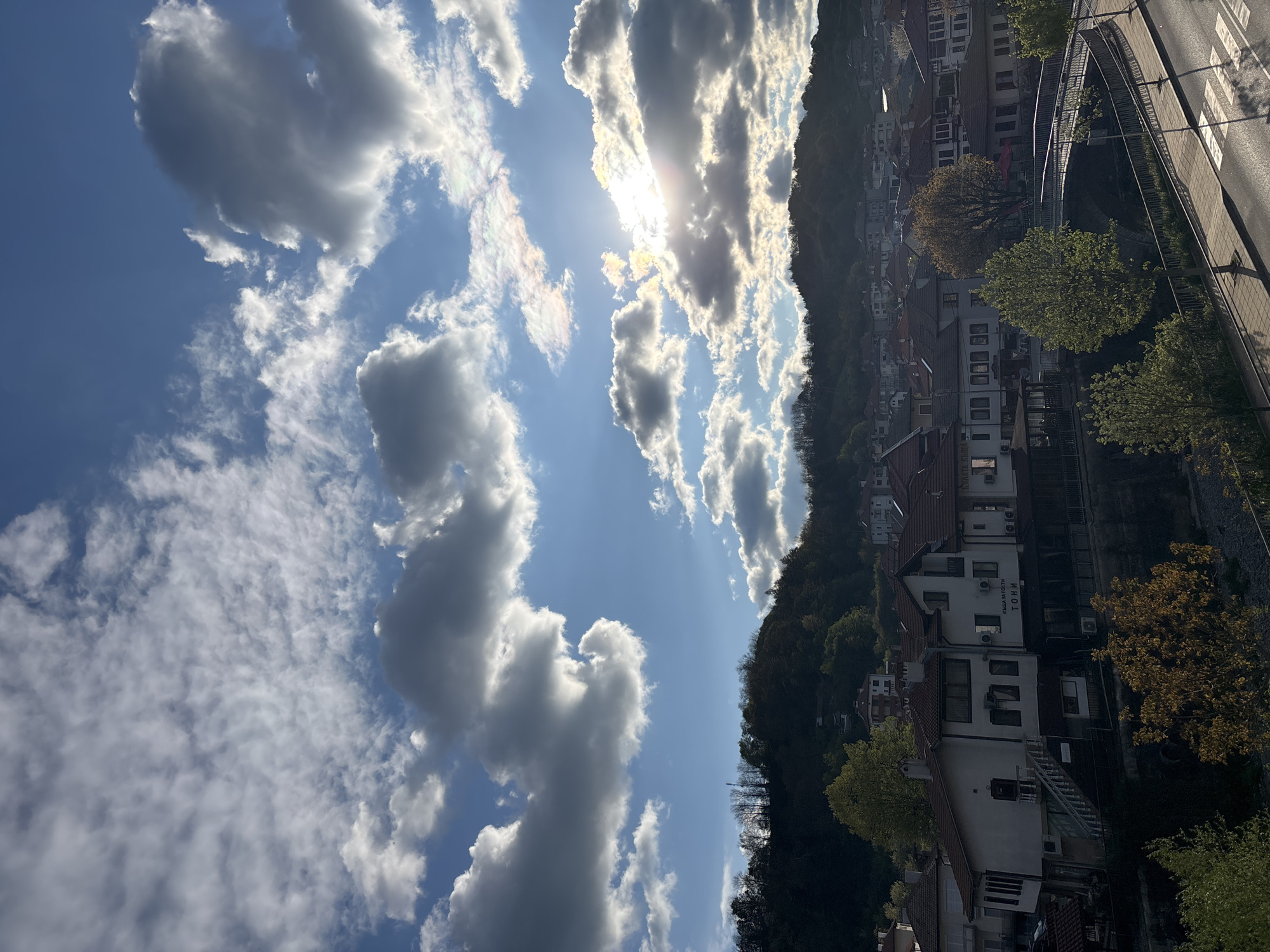 The image shows a daytime view of Zlatograd, Bulgaria. The sky is partly cloudy with bright sunlight. White cumulus clouds dominate the left side of the image, with thinner cirrus clouds visible higher up. The sun is partially obscured by the clouds. On the right, a densely wooded hillside is visible, dotted with houses and buildings. A road runs along the edge of the image. The buildings are a mix of white structures with red-tiled roofs. Trees are visible near the road and amongst the buildings. The overall impression is of a sunny day in a hilly, built-up area.