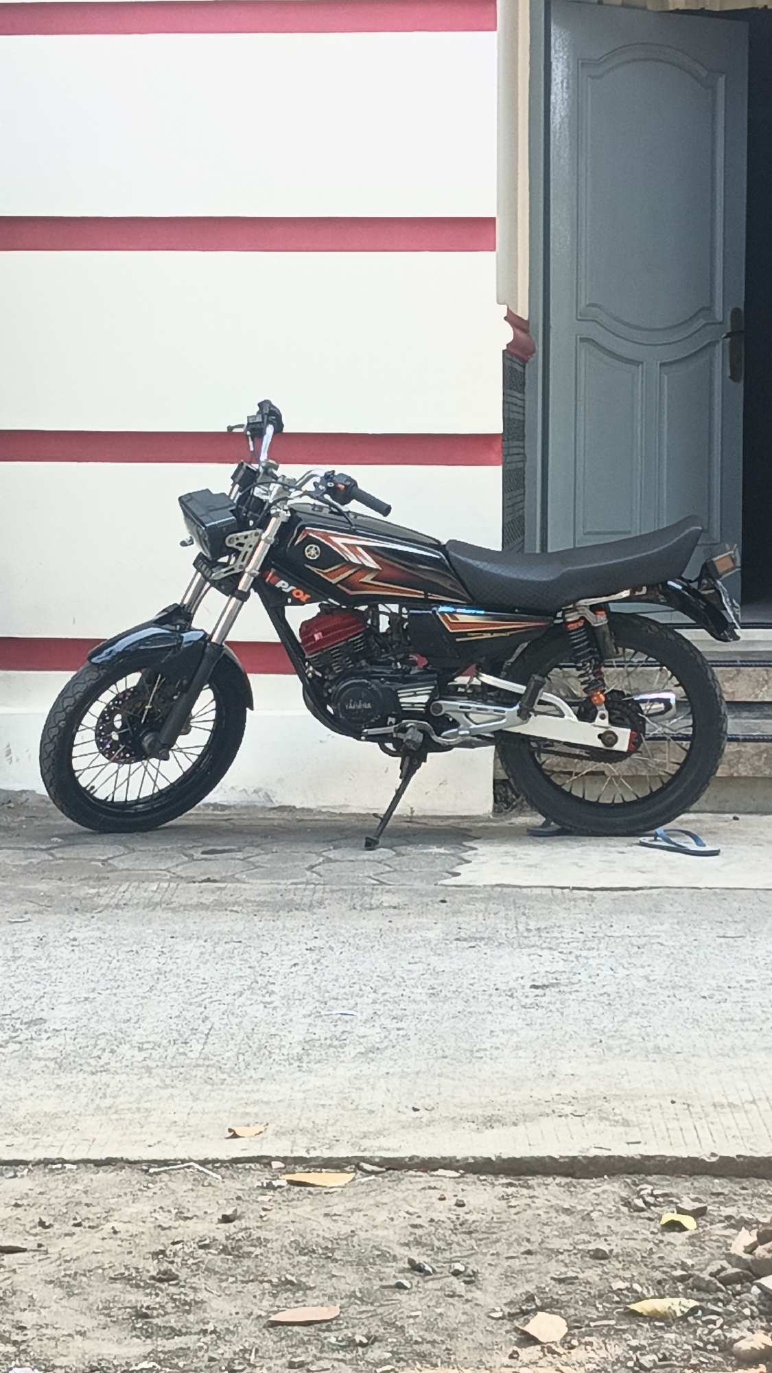 A parked black Yamaha motorcycle is positioned on a concrete surface in front of a building with a white wall adorned with two horizontal red stripes. To the right of the motorcycle, a light blue door with decorative paneling is slightly ajar, revealing a dark interior. The motorcycle features red accents on its engine and orange springs on its rear suspension. The front of the motorcycle has a black fender and a black headlight assembly. A kickstand supports the motorcycle, placing it at a slight angle. In the lower right corner of the image, a pair of blue flip-flops rests on the ground. The overall scene suggests an outdoor setting in a residential or commercial area.