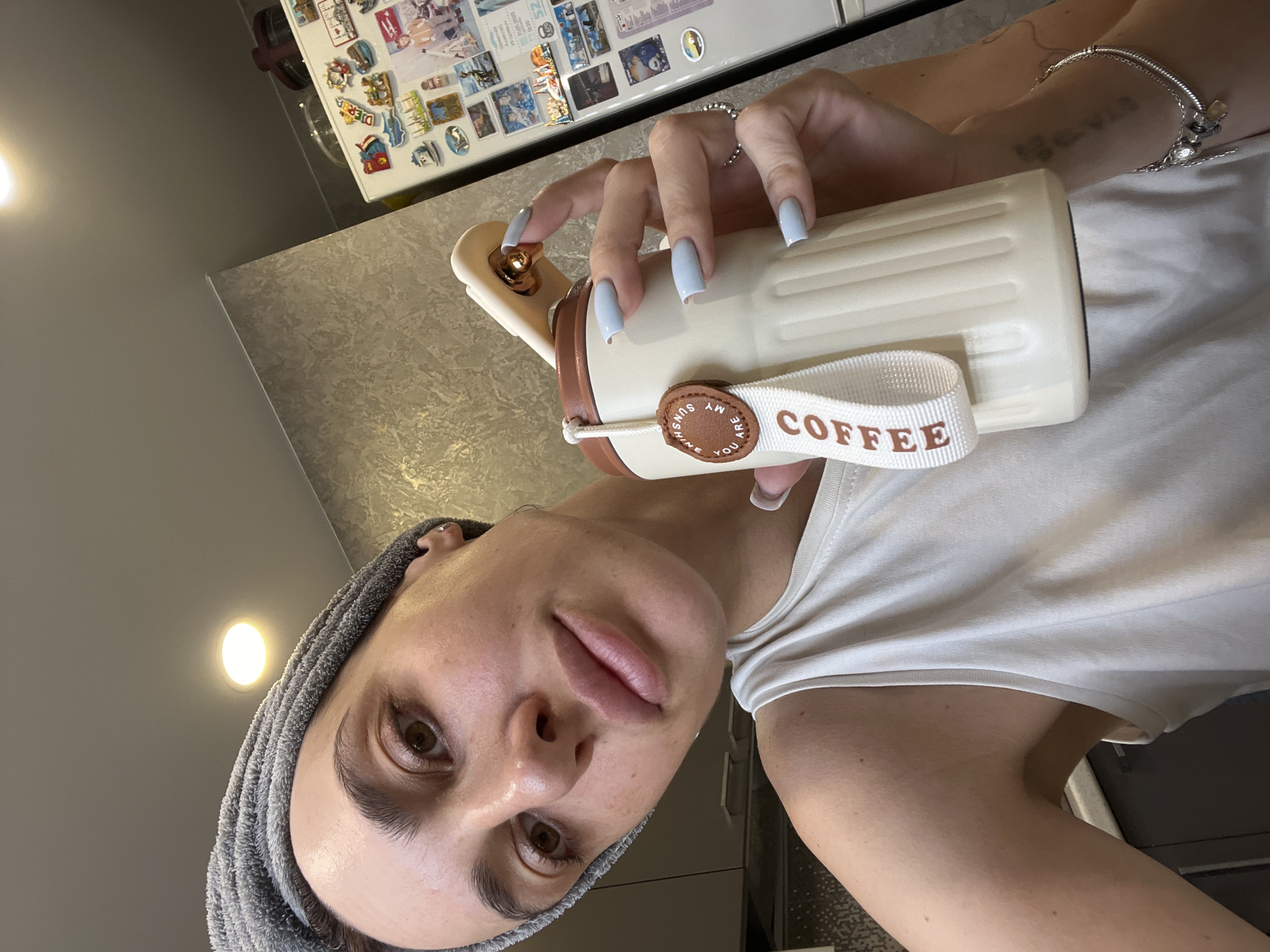 A woman with a grey towel wrapped around her head and wearing a white tank top is holding a beige tumbler with a white strap that says "COFFEE". She is indoors, possibly in a kitchen, as a refrigerator decorated with magnets is visible in the background. The lighting suggests it is daytime. The image does not provide enough information to determine the specific location within the city of Nizhny Novgorod, Russia.