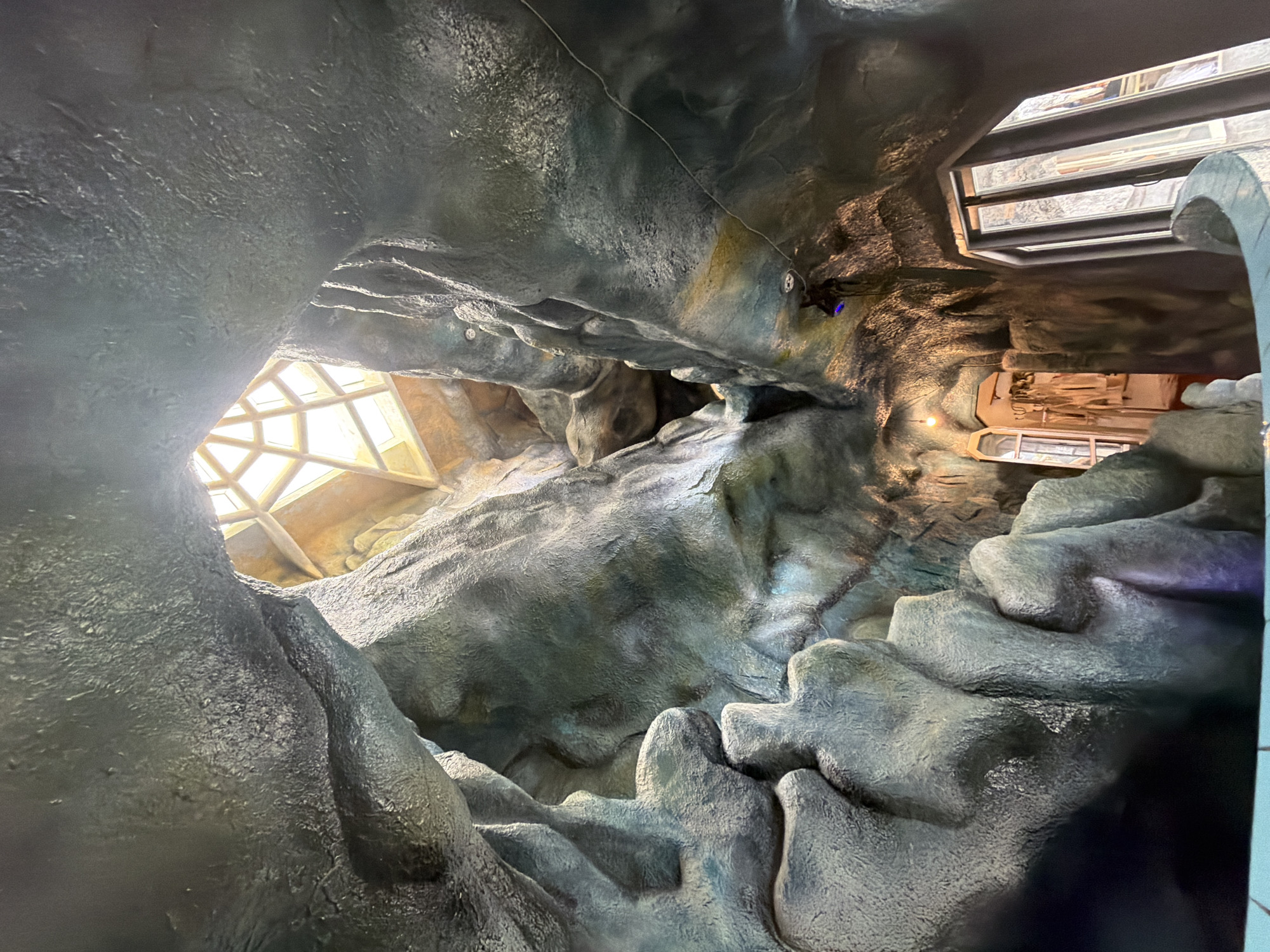 The image shows the interior of the Hang Nga Guesthouse, also known as the Crazy House, in Da Lat, Vietnam. The architecture is organic and cave-like, with winding passages and sculpted forms resembling natural rock formations. A skylight with a geometric design allows natural light to filter into the space, illuminating the textured, rough surfaces of the walls and ceiling. Several doorways and windows are visible, leading to other parts of the building, and some are adorned with ornamental details. The overall impression is of a surreal and whimsical environment, designed to evoke a sense of being in a natural grotto or fantastical landscape.