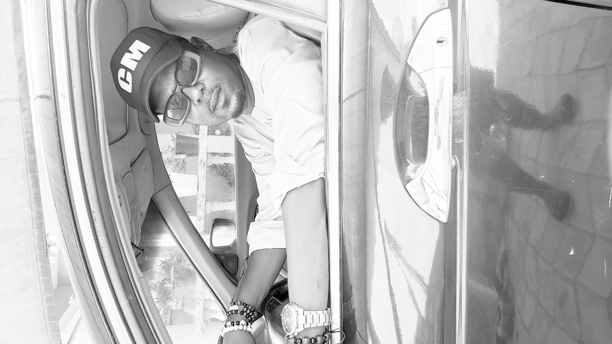 A man wearing a "CM" baseball cap and glasses leans out the window of a car. He is wearing a white shirt, bracelets, and a watch. The scene is in black and white. The background through the window shows an urban area, possibly Narayi, Nigeria, though this is speculative. The car door and handle are visible in the right of the frame.