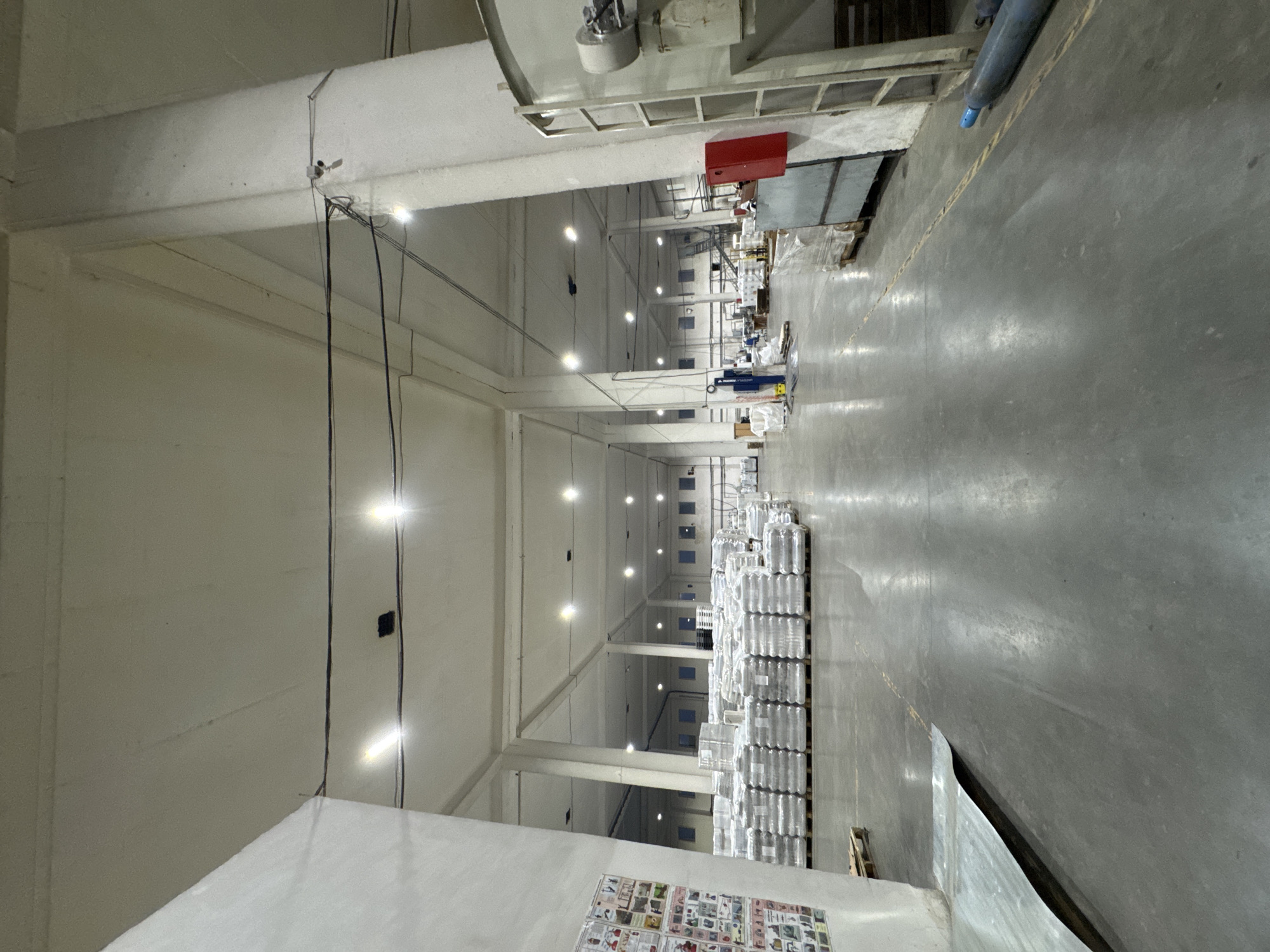 The image depicts the interior of a large warehouse facility, likely a distribution center or manufacturing plant, with a concrete floor and high ceilings. Multiple rows of palletized goods, wrapped in white plastic, are stored throughout the space. Industrial lighting fixtures are visible on the ceiling. The warehouse appears to be well-lit, though some areas are in shadow. A large, white pillar is prominently featured on the right side of the image, supporting the roof structure. Adjacent to the pillar, a red fire alarm box is mounted on a wall. Further back, a large industrial tank with a visible mechanical component is present. In the foreground, a large sheet of corrugated metal is partially covering the floor, with a yellow line marking a pathway or safety zone. The overall impression is one of an organized and functional industrial environment. There is no information in the image to suggest this location is in the city of Perm, Russia.
