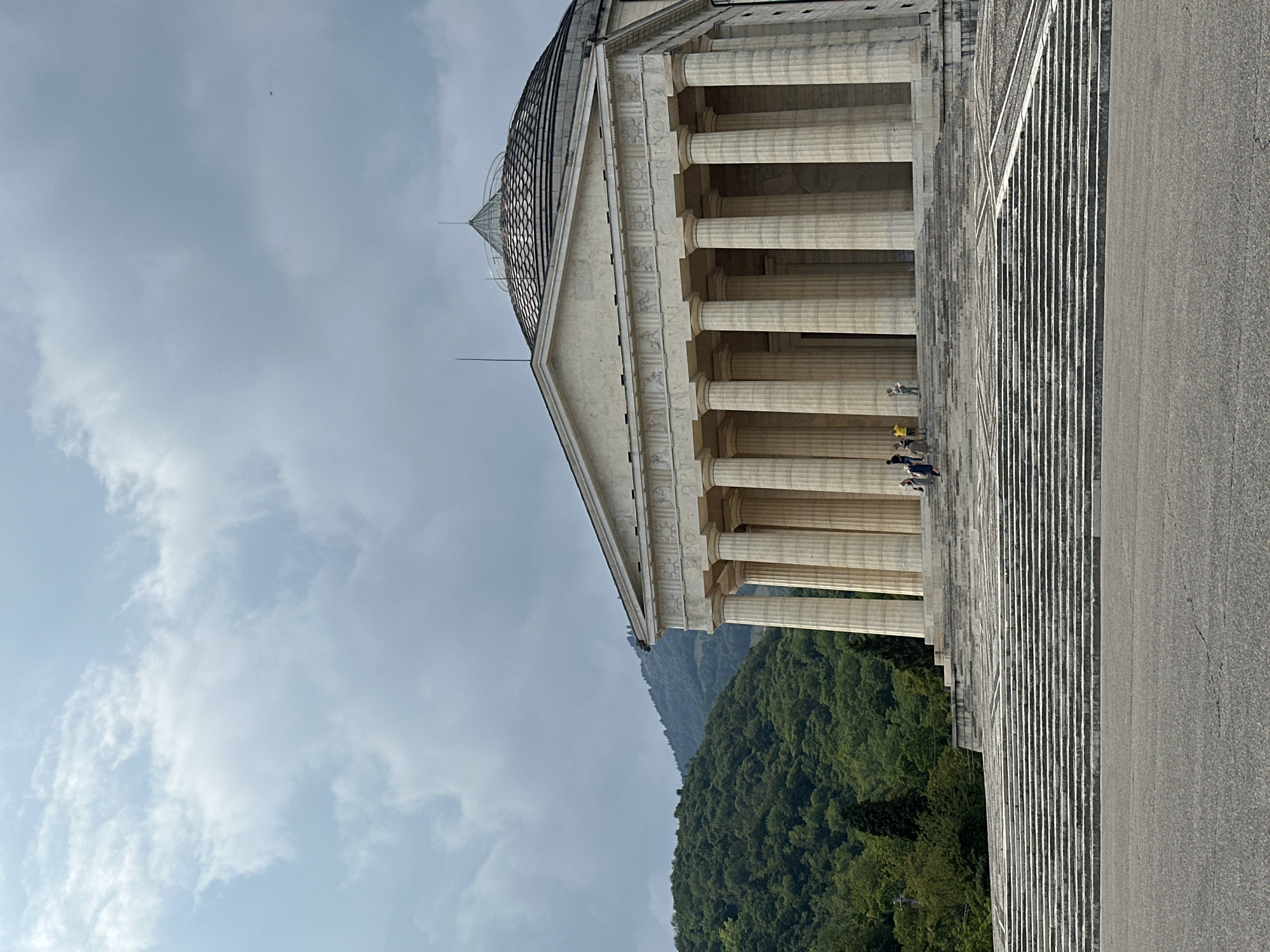 The image shows a large, Neoclassical-style building with a portico supported by numerous columns. The building is set against a backdrop of lush green hills under a cloudy sky. A wide, long staircase leads up to the entrance of the building. Several people are visible on the stairs and at the entrance, appearing to be tourists. The architecture suggests a historical monument or a public building. The location is identified as the Temple of Antonio Canova in Possagno, near Crespano del Grappa, in the province of Treviso, Italy. The building's design, with its prominent dome and classical facade, is characteristic of the works of Antonio Canova, an influential Italian sculptor. The surrounding landscape indicates a rural or semi-rural setting, likely in the pre-alpine region of Veneto.