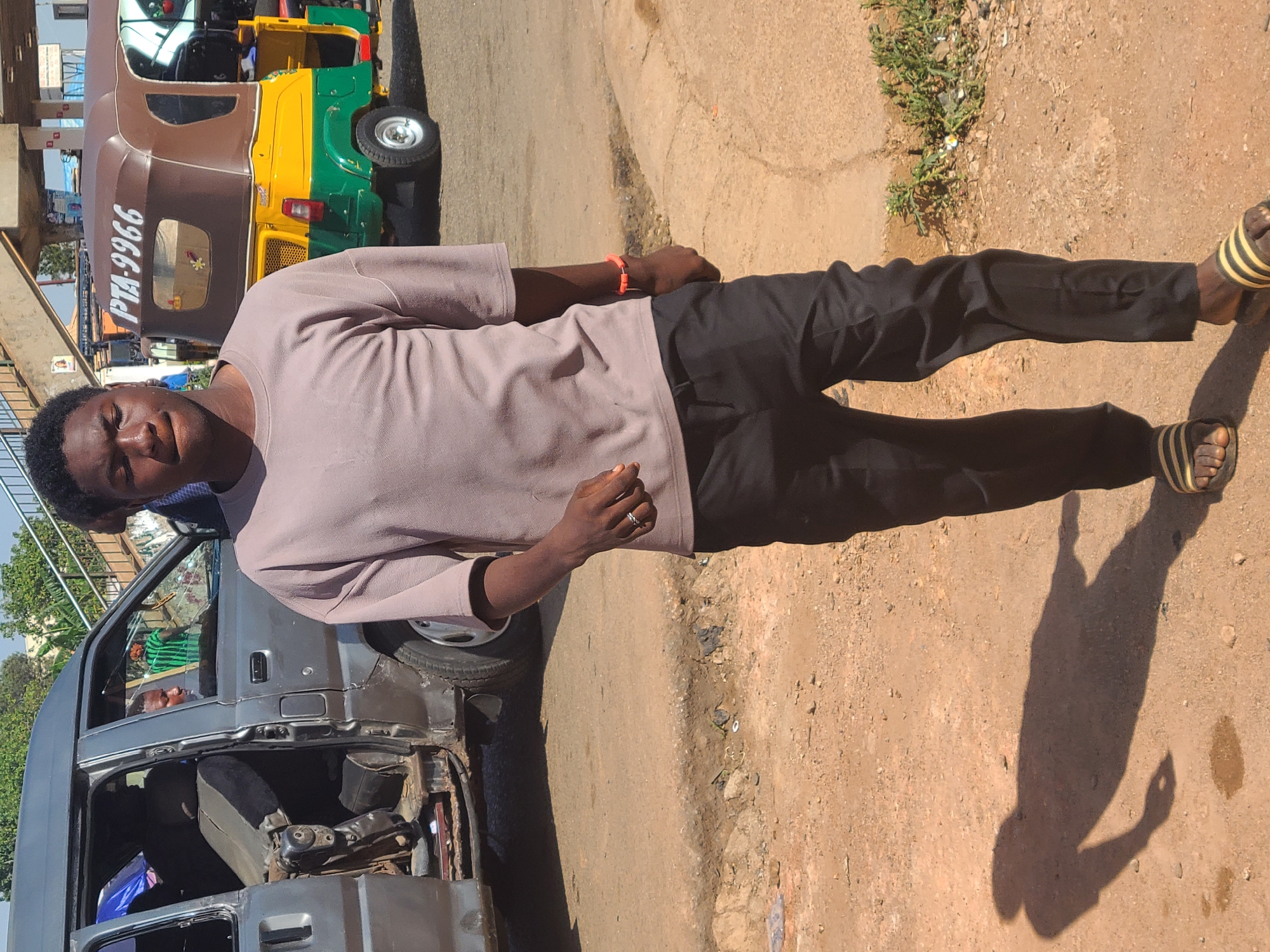 A man stands on a street in Gura Top, Nigeria. He wears a light brown t-shirt, black pants, and striped sandals. He also wears a red bracelet on his right wrist. Behind him is a grey van with a damaged side and an auto rickshaw. The ground is dirt and concrete.