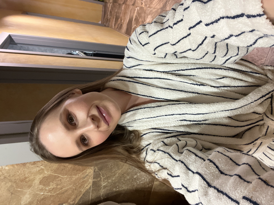 A female individual is depicted in a medium close-up, facing the camera directly, appearing to take a selfie. She has light brown hair, brown eyes, and is wearing subtle facial makeup. Her lips are slightly parted in a faint smile. She is dressed in a white terry cloth bathrobe with dark blue vertical stripes, secured at the waist. The background features two distinct areas: to the left, a wall clad with large, light brown marble-patterned tiles; to the right, a hallway with light yellow-orange painted walls. Two doorways with grey frames are visible further down the hallway on the right. The floor in the background on the right is covered with reddish-brown patterned tiles. The scene is located in a building in Khimki, Russia.