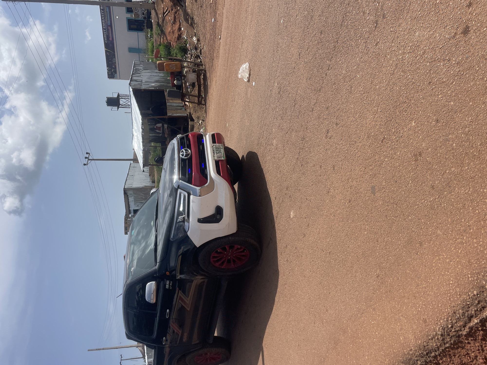 The image shows a black and white Toyota Hilux pickup truck parked on a brown, paved road. The truck has red rims and a license plate that reads NA261B02. In the background, there is a building with a sign that reads "CHRIST EMBASSY BENIN." To the left of the truck, there are some makeshift structures constructed with corrugated metal. Utility poles and wires are visible in the sky. A piece of white trash is on the ground near the truck. This scene is likely located in Durimi, Nigeria, as specified in the prompt.