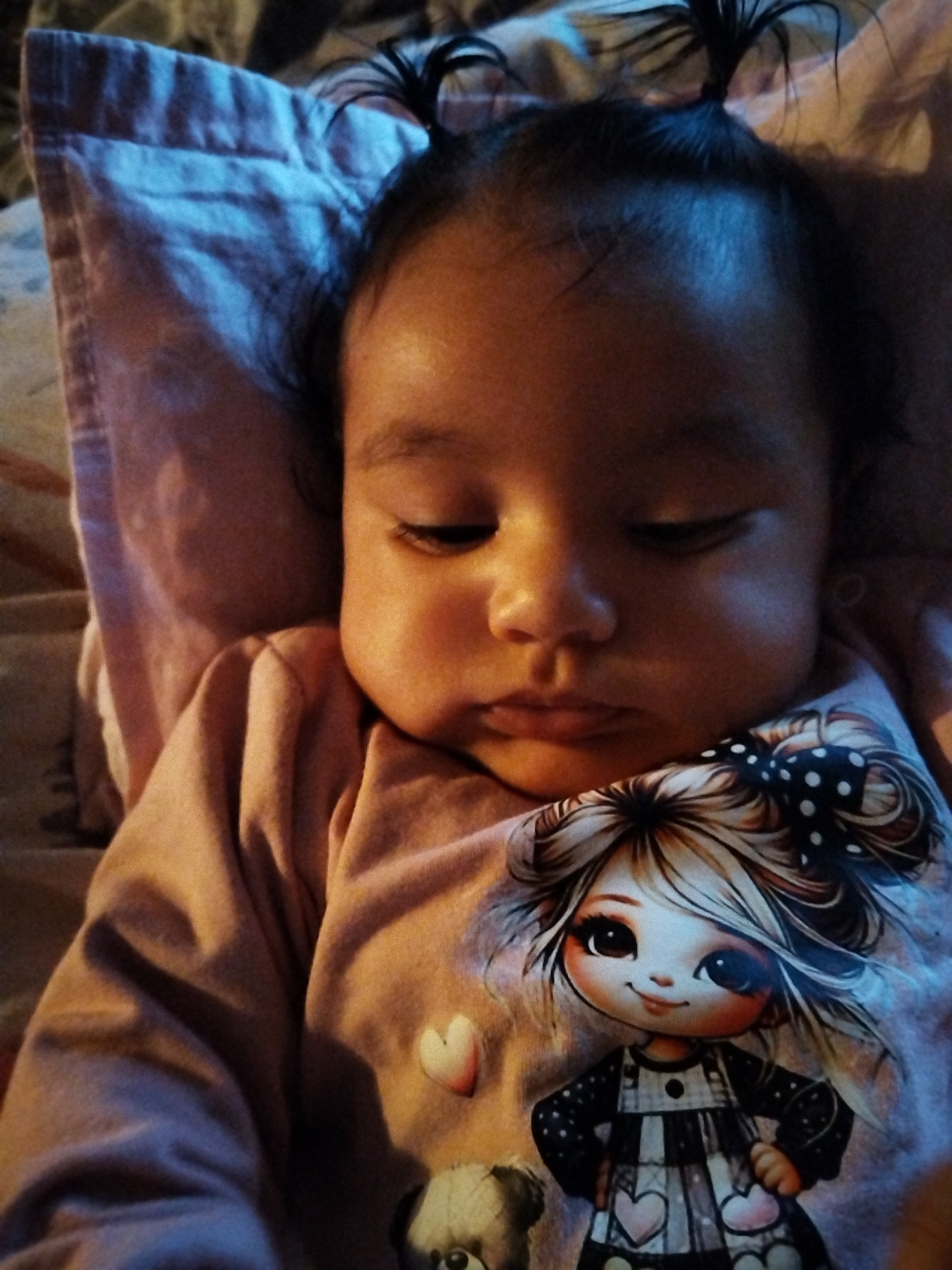 A baby is lying on a pillow. The baby has two small ponytails, dark hair, and appears to be resting or sleeping. The baby is wearing a pink long-sleeved shirt with a graphic of a cartoon girl on it. The scene appears to be indoors, possibly in a home. The specific location is unknown.