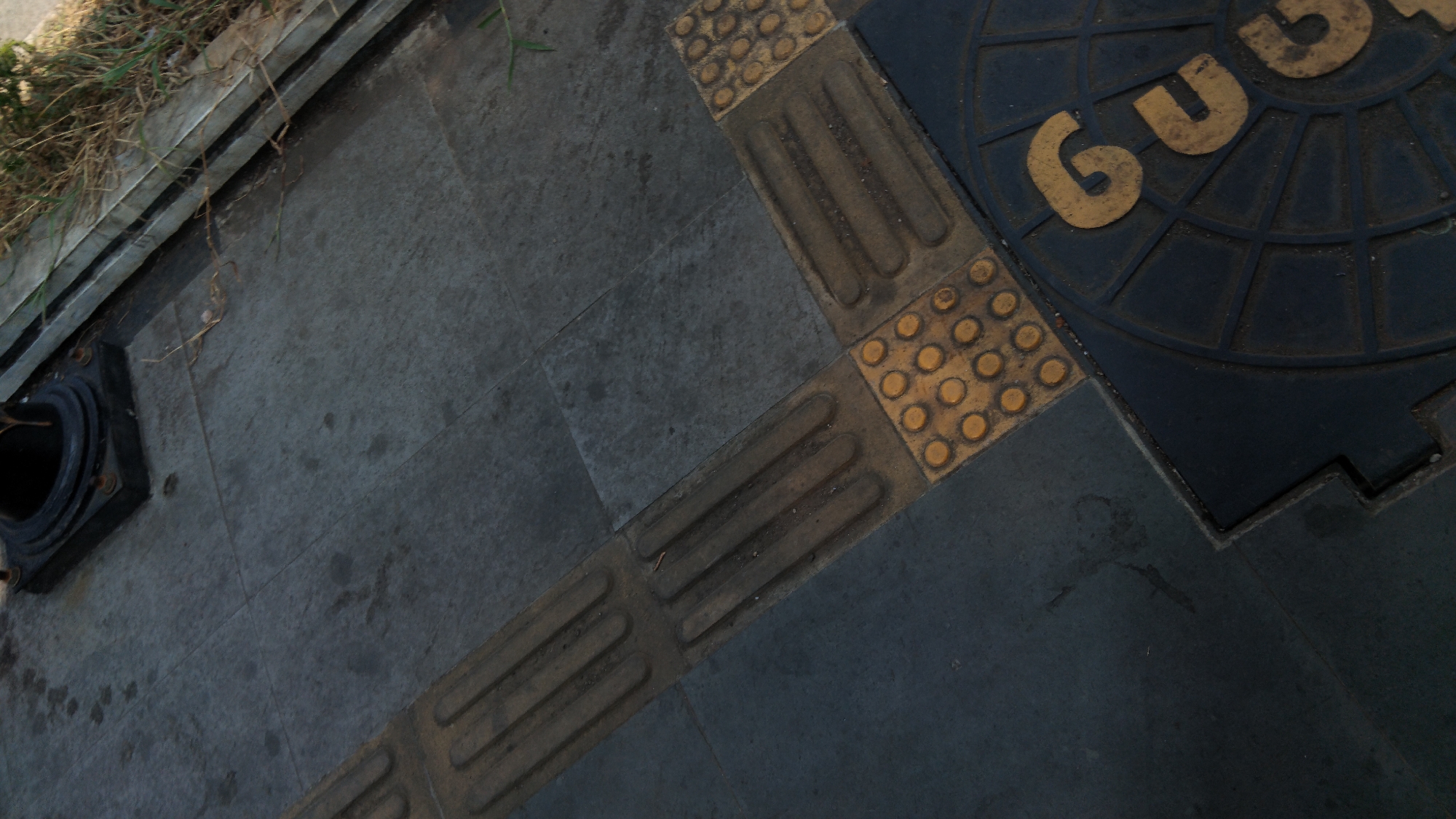This is a close-up, high-angle shot of a tactile paving section on a sidewalk in Batang, Indonesia. The paving consists of textured yellow tiles designed to warn visually impaired pedestrians. Specifically, there are rows of raised bars on some tiles and raised circular dots on others. These tactile tiles are laid on a dark gray paved surface. In the lower right corner of the image, a dark circular manhole cover is visible, with the letters "GU" in yellow prominently displayed on its surface. The surrounding environment shows some dry grass and debris near a metallic barrier, suggesting an outdoor urban setting. The lighting is natural, casting subtle shadows.