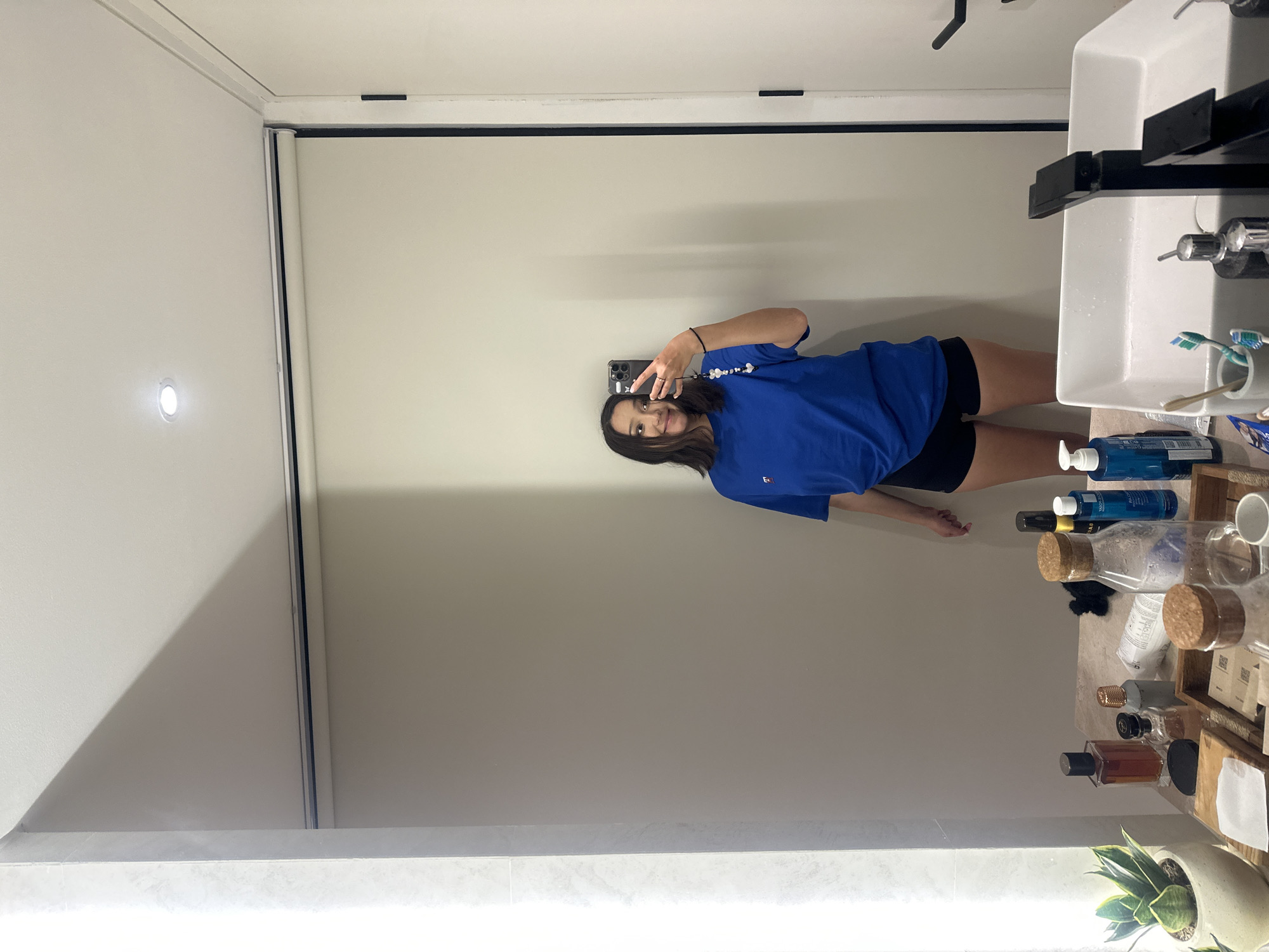A young woman is taking a mirror selfie in a well-lit bathroom, possibly in North Kuta, Indonesia. She is wearing a bright blue t-shirt and black shorts, and her left hand is raised, holding a smartphone. The phone is attached to a beaded keychain with heart-shaped charms. She is smiling and looking at her reflection.
The bathroom features a modern white sink with a black faucet, and various toiletries are arranged on the counter, including bottles of lotion and soap, toothbrushes in a holder, and what appear to be perfumes. A snake plant in a light-colored pot sits to the left of the sink. A large mirror dominates the frame, reflecting the woman and the bathroom's contents. The wall behind her is a neutral beige, with a roller blind partially visible at the top, suggesting a window or a light source. A vertical strip of light illuminates the left side of the mirror.