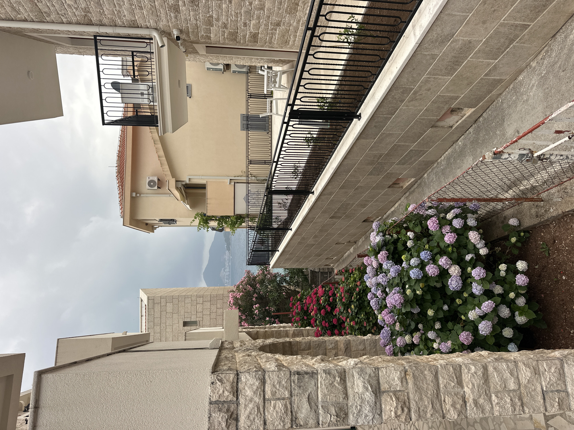 The image captures a picturesque outdoor pathway or alleyway winding between stone and stucco buildings, indicative of a Mediterranean or European setting like Pržno, Montenegro. The perspective looks along the path, with buildings framing both sides and a distant mountainous landscape visible in the background under an overcast sky.
On the right side, a vibrant display of flowering shrubs, primarily hydrangeas, lines an elevated section. The hydrangeas showcase a beautiful mix of purple, pink, and white blooms. This section is bordered by a robust stone wall made of roughly cut blocks and topped with black wrought-iron railings. Beyond this, an older, worn chain-link fence is visible on the lower ground, adjacent to a concrete surface.
On the left, a light-colored stucco building features balconies with black wrought-iron railings and an external air conditioning unit. Across the path, the building on the right also has balconies, one of which displays white outdoor furniture (a table and chairs) and another air conditioning unit. The walls of some buildings feature light-toned stone cladding, complementing the stucco.
The ground is paved with rectangular stone tiles on the elevated sections and a rougher, darker surface on the lower path. The soft, diffused light suggests a daytime scene, possibly late morning or afternoon, with no harsh shadows due to the cloudy conditions. No people are visible, contributing to a serene and tranquil atmosphere. No discernible text is present in the image.