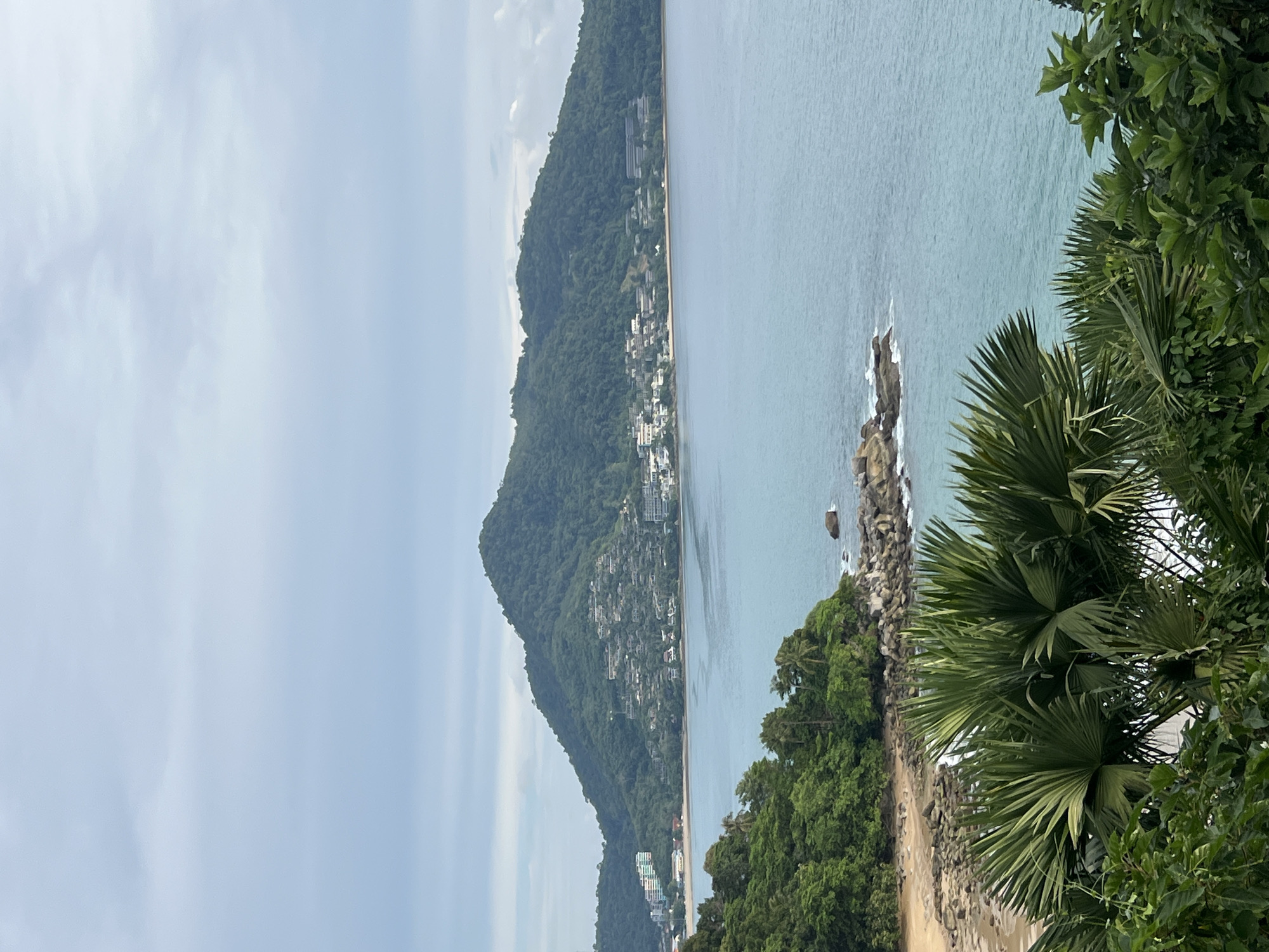 A wide shot captures a coastal landscape in Ban Bang Thao, Thailand, under an overcast sky. The image is divided into three horizontal planes. The top plane is filled with a muted blue sky with scattered white clouds. The middle plane is dominated by the serene expanse of the ocean, its surface a tranquil turquoise. Along the horizon, a lush, green mountainous island rises, dotted with clusters of buildings that appear to be hotels or residential complexes. The island slopes down to meet a sandy beach, where the ocean gently laps. In the foreground, the immediate shoreline is rocky and sparsely vegetated, with a small, sandy cove visible. The bottom plane is a dense tapestry of lush green foliage, including palm fronds, that partially obscures the view of the beach and ocean. The scene is devoid of any human presence or discernible activity, presenting a peaceful, natural vista.