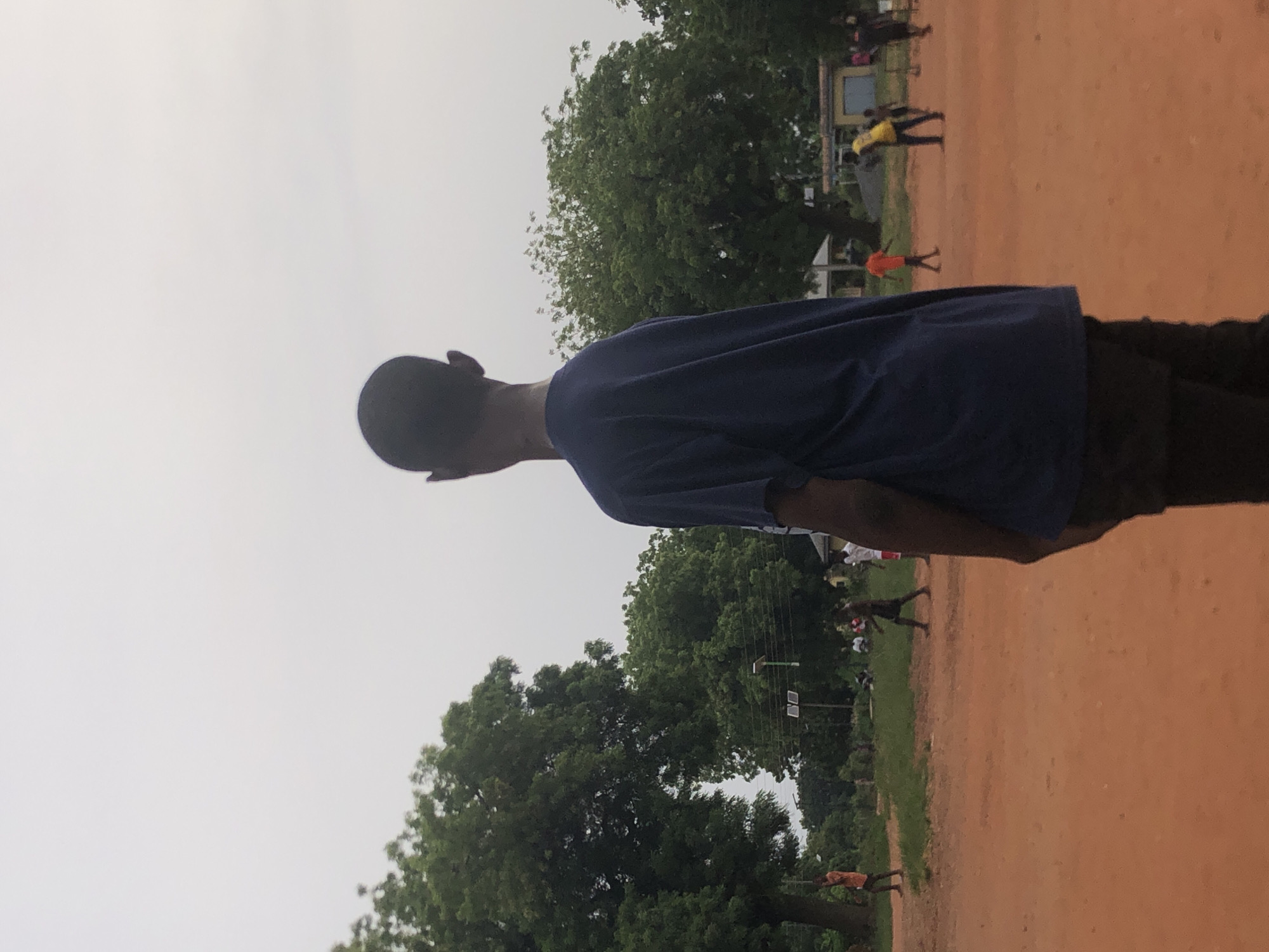 This media file captures an outdoor scene, likely in Nsugbe, Nigeria, under an overcast sky.
The main subject, positioned in the foreground, is a young person, presumably male, with short dark hair. They are seen from behind, wearing a navy blue short-sleeved t-shirt and dark trousers, looking towards the right side of the frame.
The setting is a large, open field covered in reddish-brown dirt or sand. The background features a line of dense, green trees, suggesting a natural or semi-rural environment. The sky is a uniform light grey, indicating an overcast day without direct sunlight.
In the mid-ground and background, several other individuals are visible. One person stands out in a bright yellow shirt and dark pants, near a light-colored building or structure. Further back, a smaller figure in an orange top is present. Other people are scattered across the field and near the treeline, appearing to be walking, standing, or engaging in casual activities. Utility poles or power lines are faintly visible among the trees.
The image depicts a casual outdoor gathering or daily life in an open community space, possibly a schoolyard or a local field. The time of day cannot be precisely determined but appears to be either early morning or late afternoon due to the diffuse light, or simply a cloudy day. There is no visible text in the image.