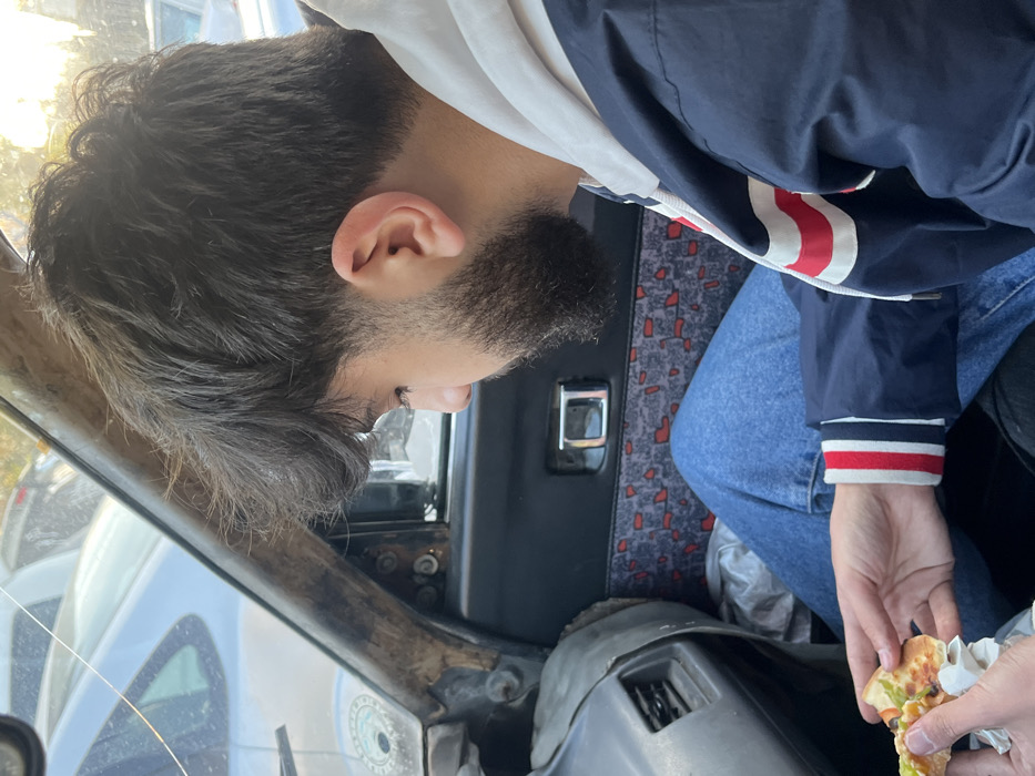 Within the Kafr Sousa Municipality, Syria, a male individual is seated inside a vehicle, observed from the chest up in profile, looking downwards. He has dark, unkempt hair and a dark beard, and is wearing a white hooded sweatshirt beneath a navy blue jacket with white and red striped cuffs and accents, along with blue denim jeans. His hands hold a partially consumed slice of pizza, wrapped in white paper, featuring visible green pepper, black olive, and cheese toppings.
The immediate environment is the interior of a vehicle, displaying signs of wear and age. The left A-pillar is visible, appearing worn and possibly damaged, next to a cracked windshield through which other vehicles and daylight are discernible. A dashboard or side panel section to the left includes an air vent. The vehicle's door panel to the right features a patterned fabric insert with red and light blue elements and a chrome door handle. The external environment, seen through the vehicle's windows, shows other parked or stationary vehicles, suggesting an outdoor urban setting.