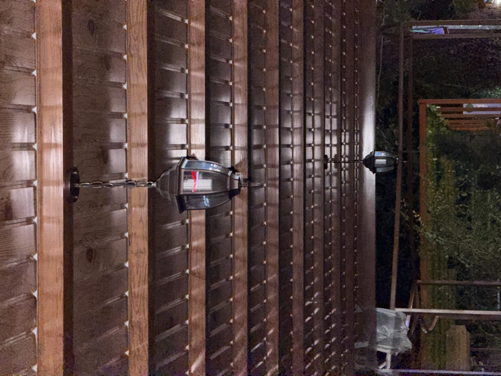 An outdoor nighttime shot shows a wooden pergola with a series of hanging lanterns. The pergola's roof is made of horizontal wooden beams with angled slats underneath, creating a textured, shaded effect. Two lanterns are visible. The upper lantern, closer to the camera, is a black metal fixture with glass panels, one of which displays a red and blue light. A second, similar lantern is positioned further back and lower down. Below the pergola, dimly lit foliage and structures can be seen. The scene appears to be in a garden or patio area.