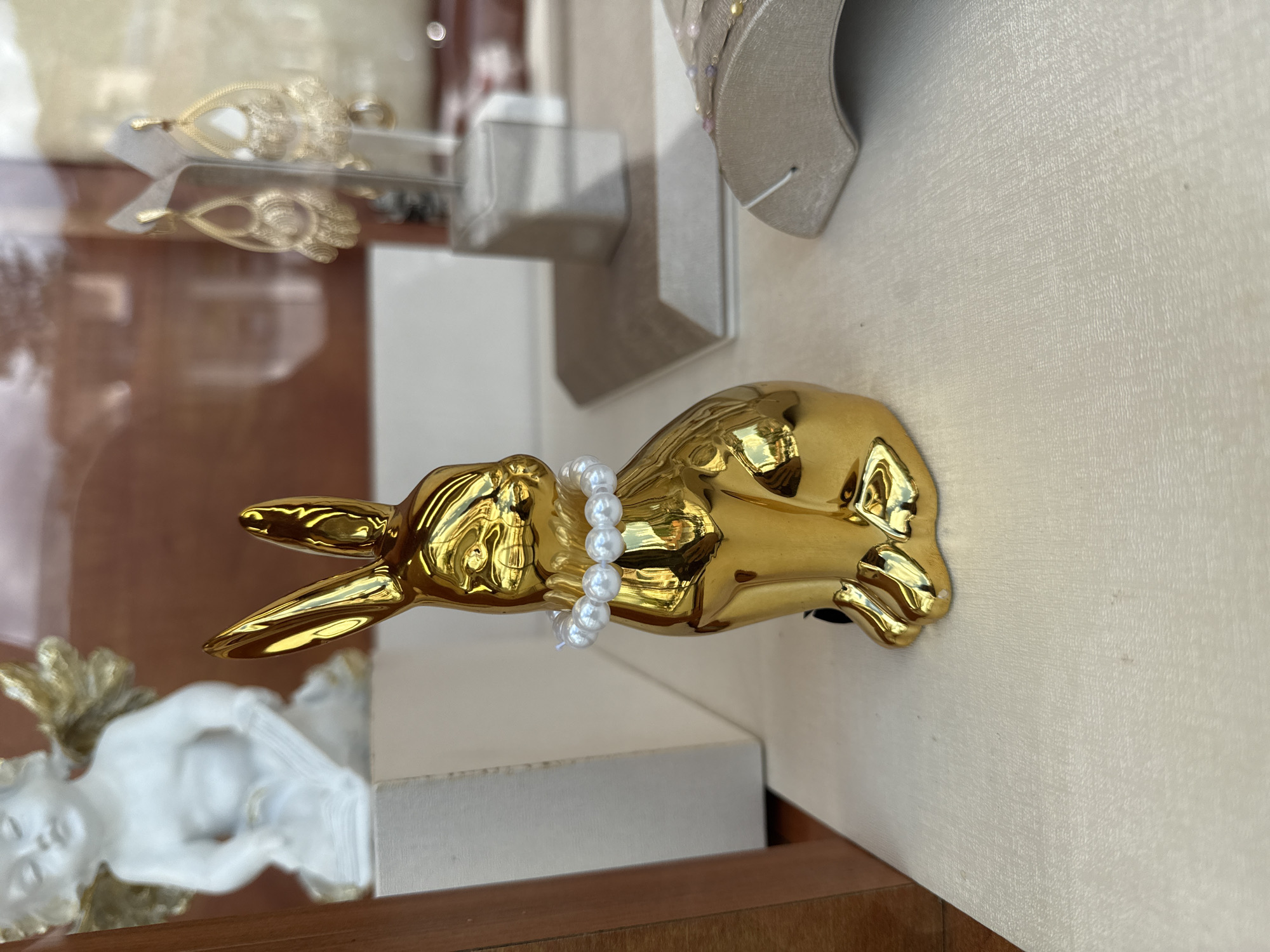 A golden rabbit statuette with a string of pearls around its neck lies on its side on a surface. The scene appears to be inside a shop or display case, as reflected objects are visible in the background. Other items are also present, including a white figurine and a neutral-colored object in the upper right. The location of the scene is Veliko Tarnovo, Bulgaria.