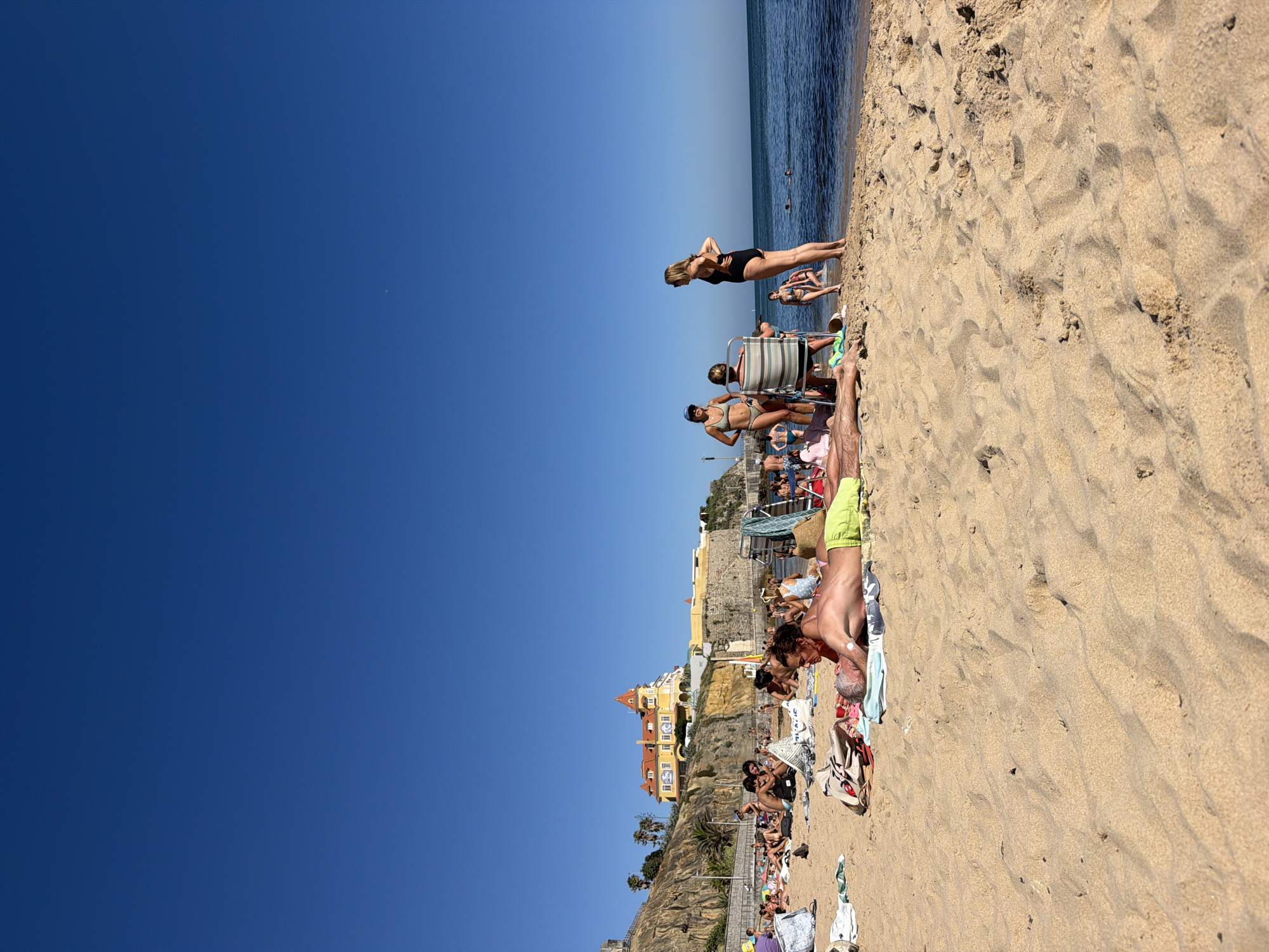 The image depicts a sun-drenched beach scene in Cascais, Portugal. The foreground is dominated by textured sand with footprints, indicating recent activity. A clear, deep blue sky stretches across the upper portion of the frame. In the mid-ground, a group of people are scattered across the sand, some lounging on towels and beach chairs, while others are in the water. The ocean is a calm blue, meeting the sandy shore.
Further back, buildings with distinctive architecture, characteristic of coastal Portugal, are visible on a cliffside overlooking the beach. These buildings are colorful, with terracotta roofs and yellow facades, suggesting a resort or town area. Some palm trees are interspersed among the buildings and on the cliffs.
The people are dressed in swimwear, engaged in typical beach activities like sunbathing and swimming. Their poses and the overall atmosphere suggest a leisurely summer day. The sun is high in the sky, indicating it is likely midday or early afternoon. The weather appears to be warm and clear, with no clouds visible. The mood is relaxed and vacation-like. There is no visible text in the image.
