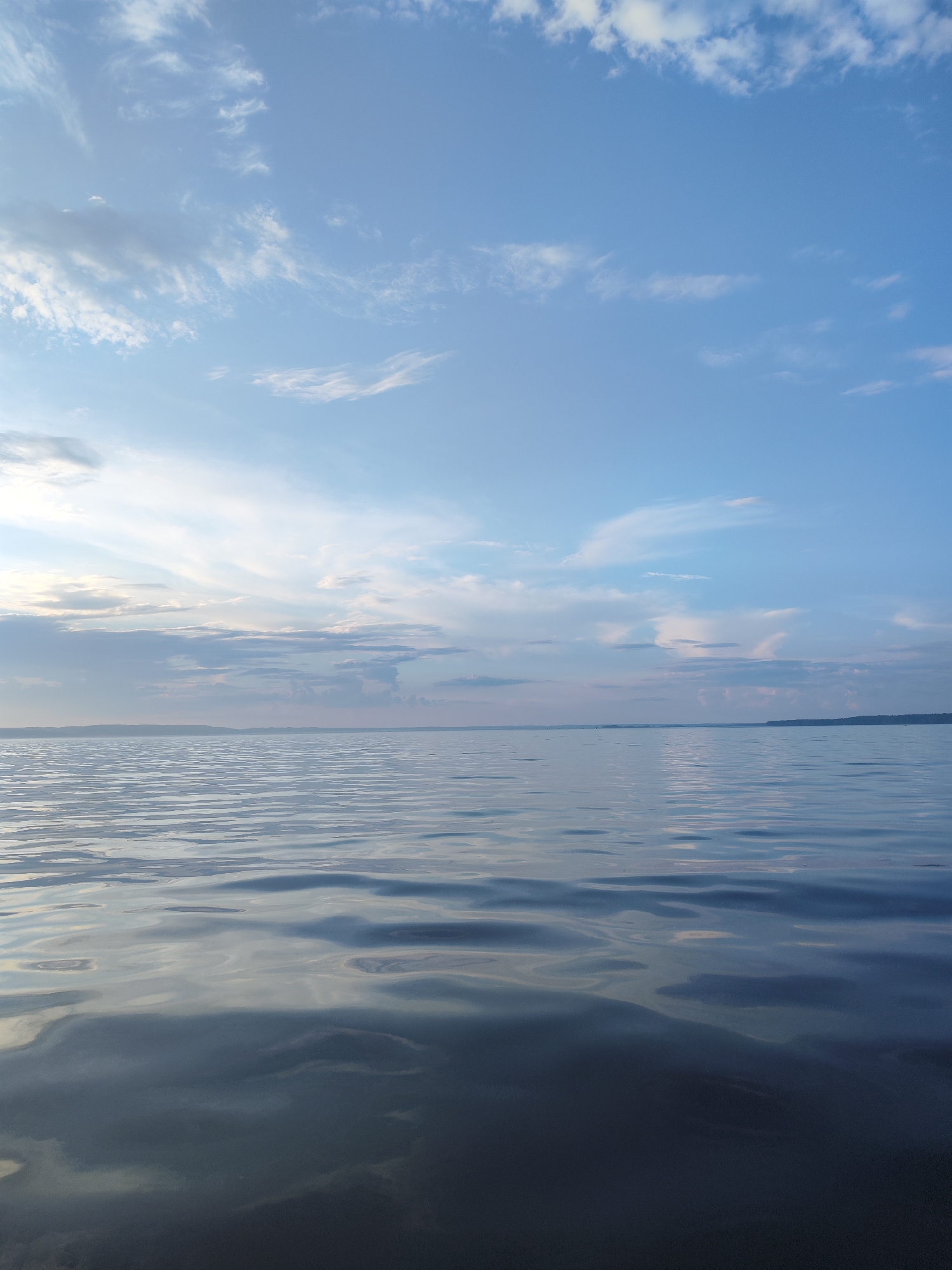 A vertical shot captures a serene body of water and a cloudy sky. The water, in the foreground, exhibits gentle ripples, reflecting the pale hues of the sky. The horizon line, a thin strip of land, separates the water from the sky. The sky above is a soft blue, adorned with scattered wispy clouds, some tinged with pink and orange, suggesting either sunrise or sunset. The overall scene evokes a sense of tranquility and natural beauty.
While the image depicts a body of water and sky, it does not contain any elements that would allow for a specific identification of its location within the city of Niva, Russia, or any other identifiable landmarks. There are no people, structures, or specific geographical features to pinpoint a precise location.