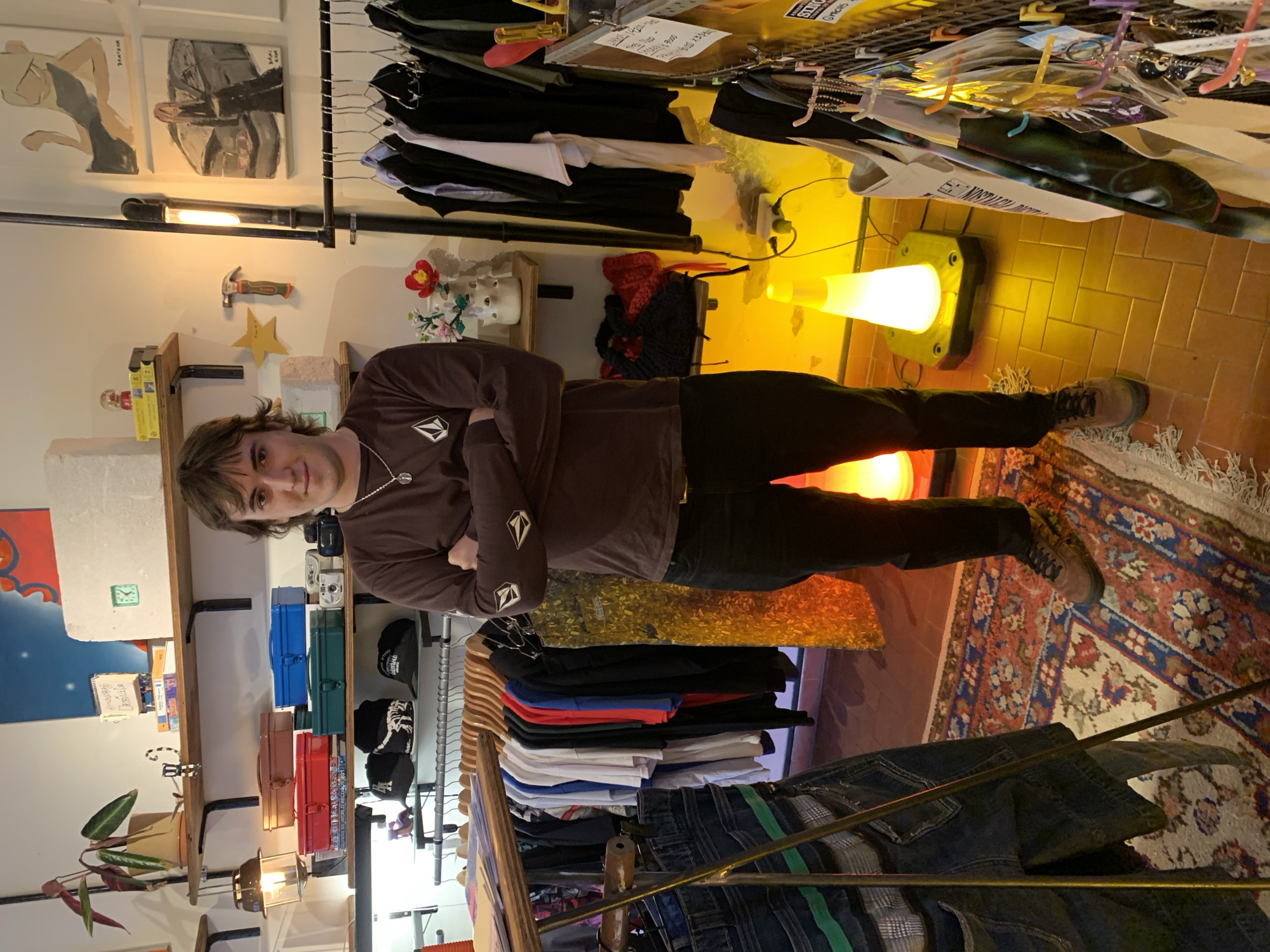 The image shows a man standing with his arms crossed in what appears to be a retail store. He is wearing a brown long-sleeved shirt, dark pants, and boots. The store has clothing racks with various garments including shirts and jeans. Shelves display items like metal boxes and hats. The lighting in the store is a mix of natural light and artificial lighting including a yellow traffic cone lamp. A patterned rug is visible on the floor. The location is indicated by the presence of various artifacts indicative of Buenos Aires Argentina, including cultural decor, clothing and objects.