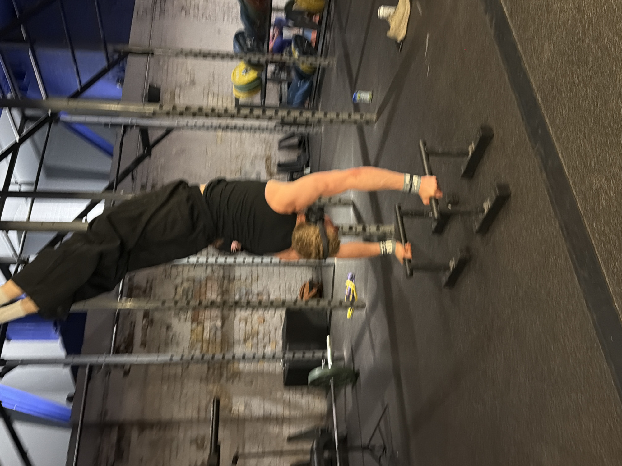A person is performing a gymnastic skill called a "planche" on parallettes in a gym. The person is wearing a black tank top and black pants, with wrist wraps on both wrists. The person's body is horizontal, supported only by their arms. The gym has a brick wall, a black floor, and a rack of weights and pull-up bars in the background. The location is likely not in London, United Kingdom.