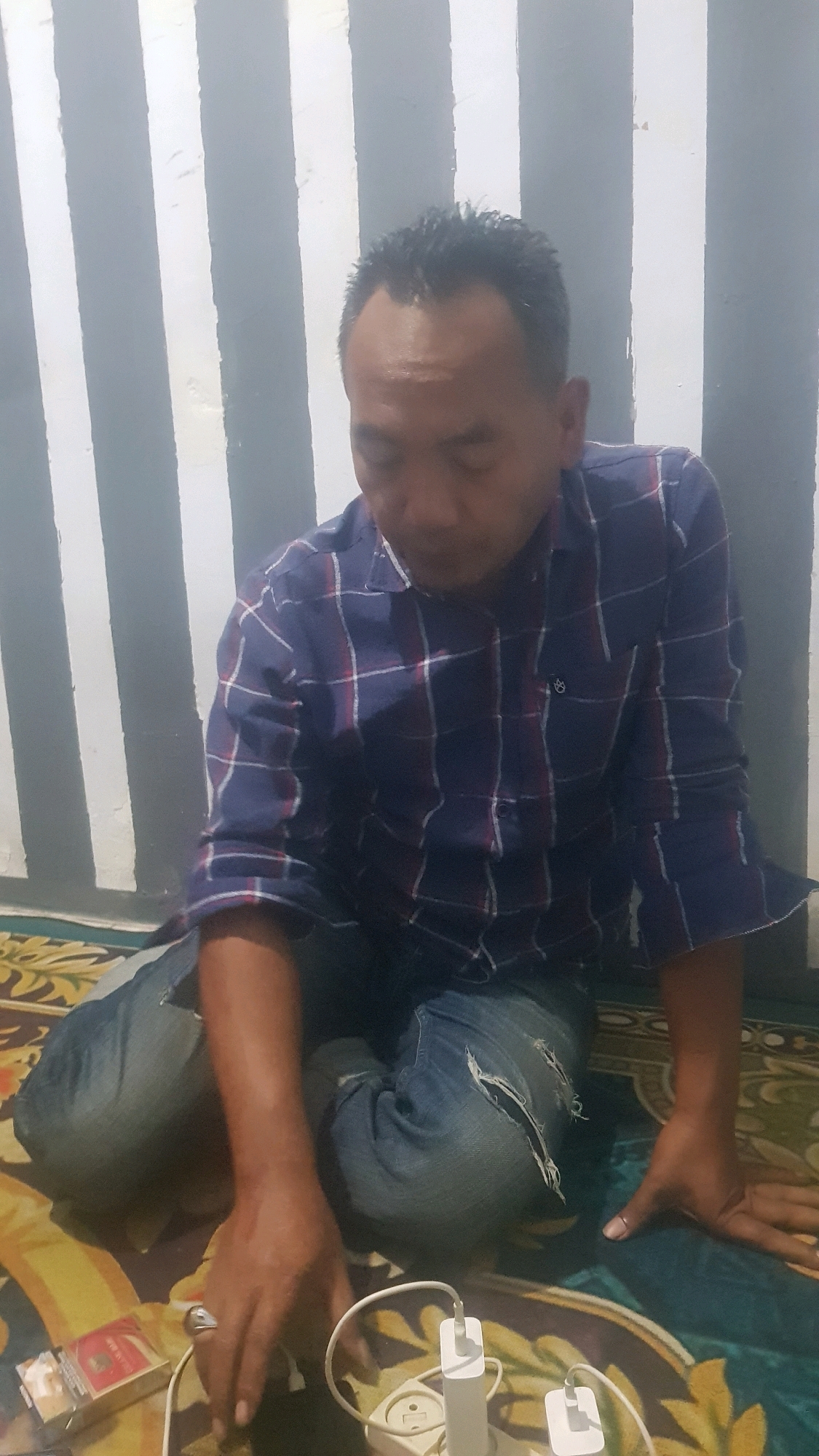 A man is sitting cross-legged on a patterned rug in what appears to be a room with a striped wall. He is wearing a blue and red plaid shirt and ripped blue jeans. His head is bowed, and his eyes are closed. A pack of cigarettes and a charging adapter with two cords are on the rug in front of him. The scene is lit by an overhead light. The location is confirmed to be Bakalan, Indonesia.