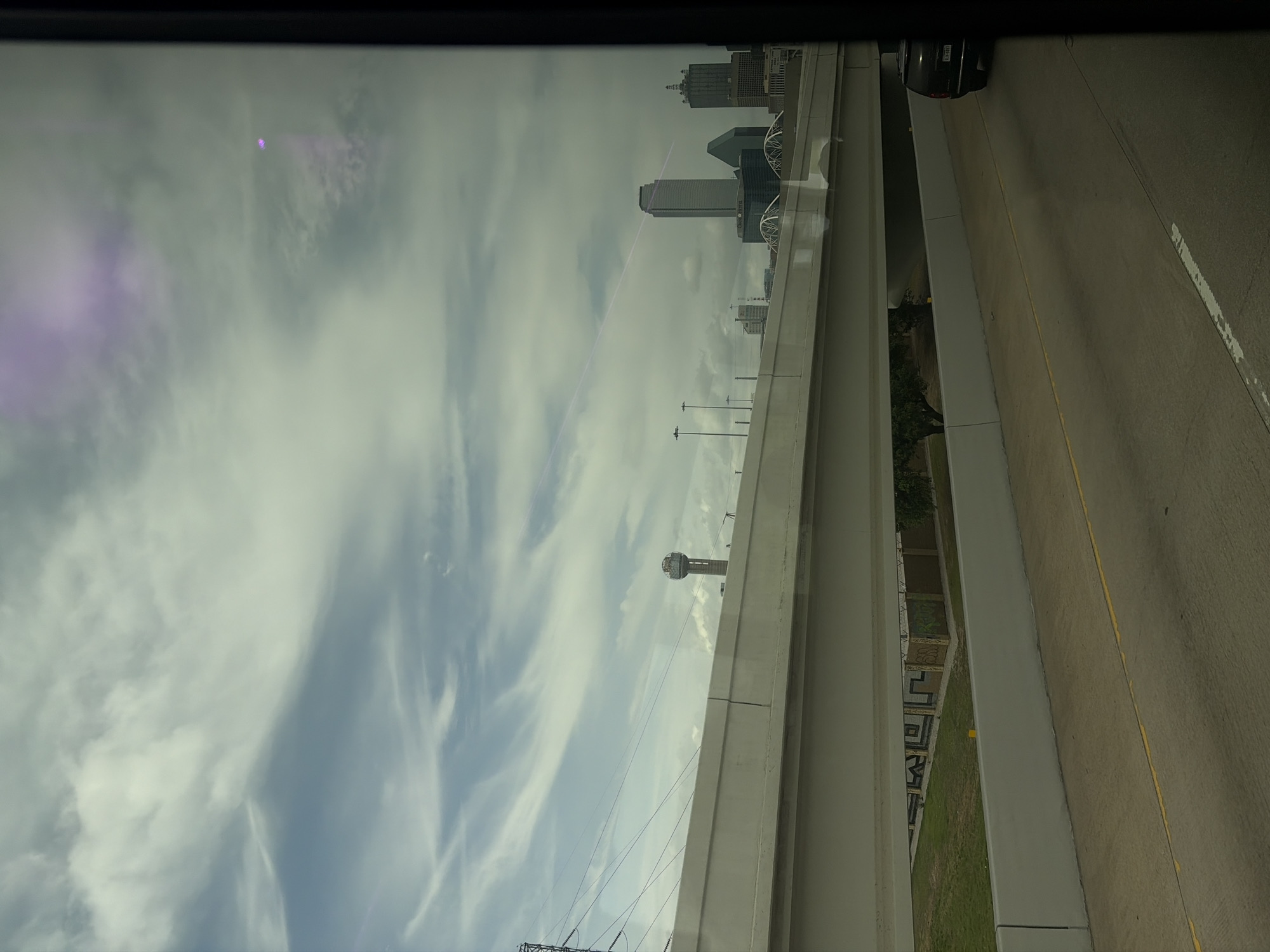 The image shows a view from an elevated position, possibly a bridge or overpass, overlooking a highway and the Dallas skyline. The skyline includes Reunion Tower. A black car is on the highway, along with a yellow dividing line. The sky is cloudy.