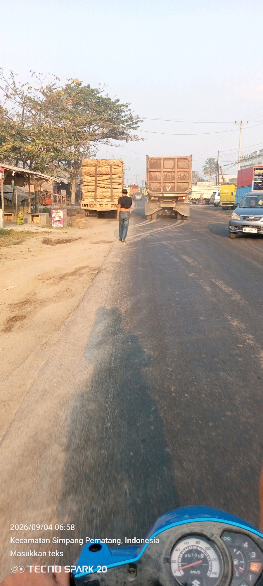 The image captures a scene on a road in Kecamatan Simpang Pematang, Indonesia, on September 4, 2026, at 06:58. A man in a black t-shirt and blue jeans walks away from the camera on the right side of the road. He is carrying a cigarette in his right hand. The road is paved and appears to be a two-lane highway. On the left side of the road, there is a dirt shoulder with a small building structure and a parked truck loaded with logs. Further down the road, a large dump truck is in motion, and other vehicles are visible in the distance. The sky is clear and bright, suggesting it is early morning. The image is captured from the perspective of a motorcycle, with a portion of the handlebar and speedometer visible in the foreground. The speedometer reads 26705.6 km/h.