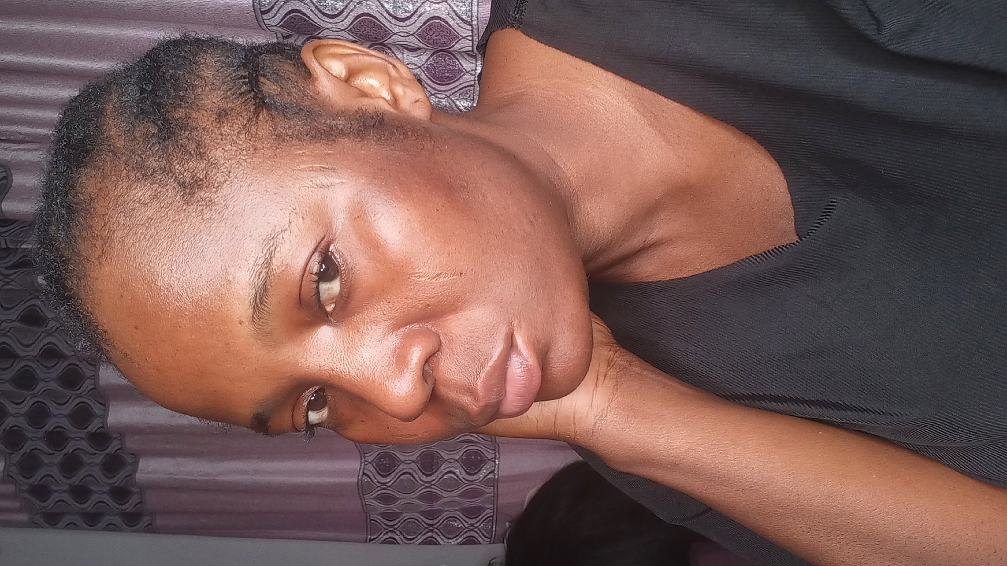 A medium shot of a young woman in a black top shows her face in profile, with her head tilted slightly to the left and her left hand resting on her jawline. Her hair is styled in short braids, and her skin is dark and smooth. The background consists of a patterned curtain in muted tones of purple and gray. The lighting is soft and diffuse, creating a warm glow on her face. The woman's expression is thoughtful, with her eyes looking slightly off-camera.