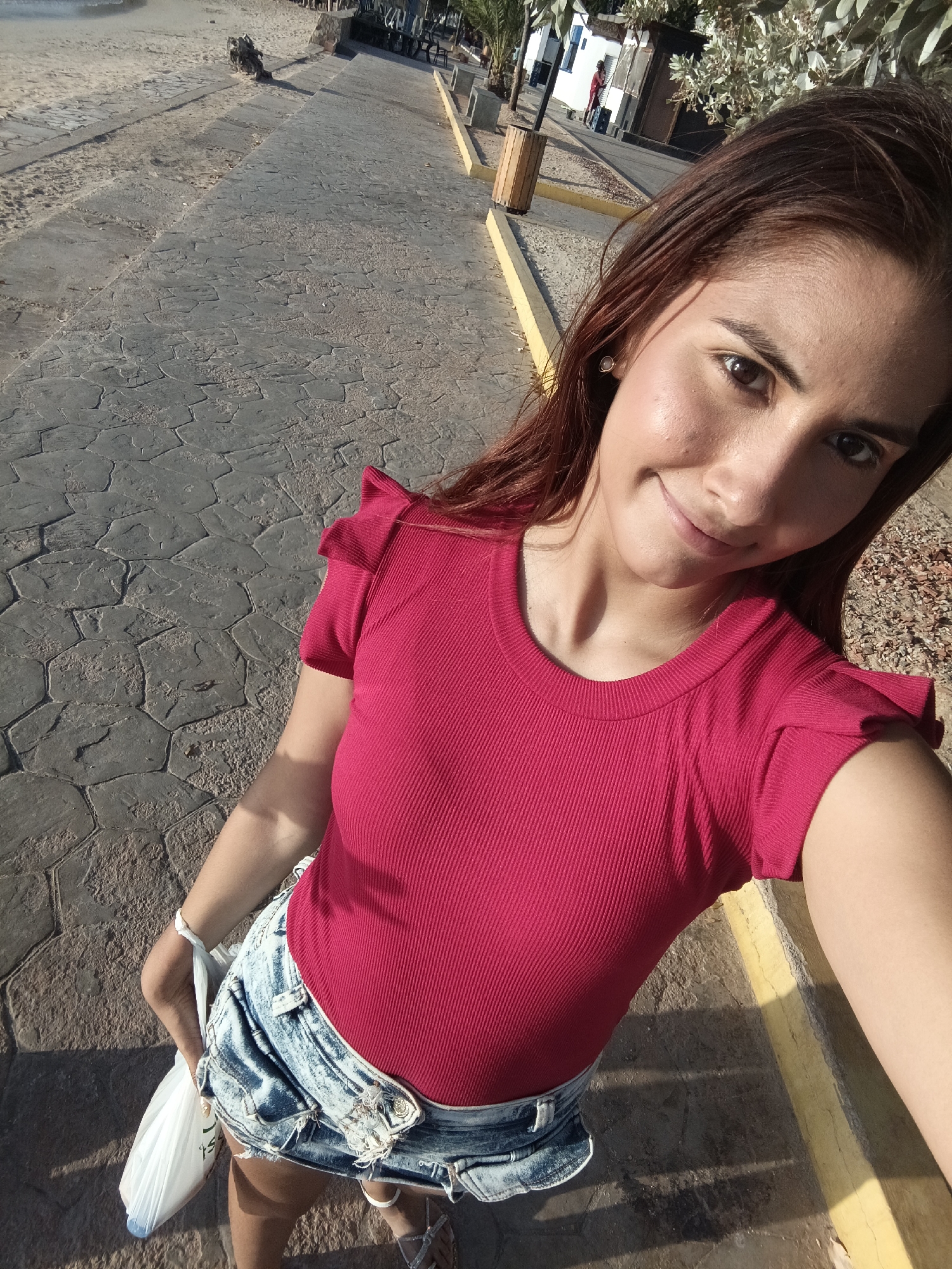 A woman is taking a selfie on a paved walkway in Juan Griego, Venezuela. The walkway is constructed of irregular, interlocking paving stones and is bordered by a low, yellow curb. In the background, there is a beach and a row of buildings, some of which are white with blue trim. The woman is wearing a red, short-sleeved shirt and denim shorts. She is holding a white plastic bag in her left hand.