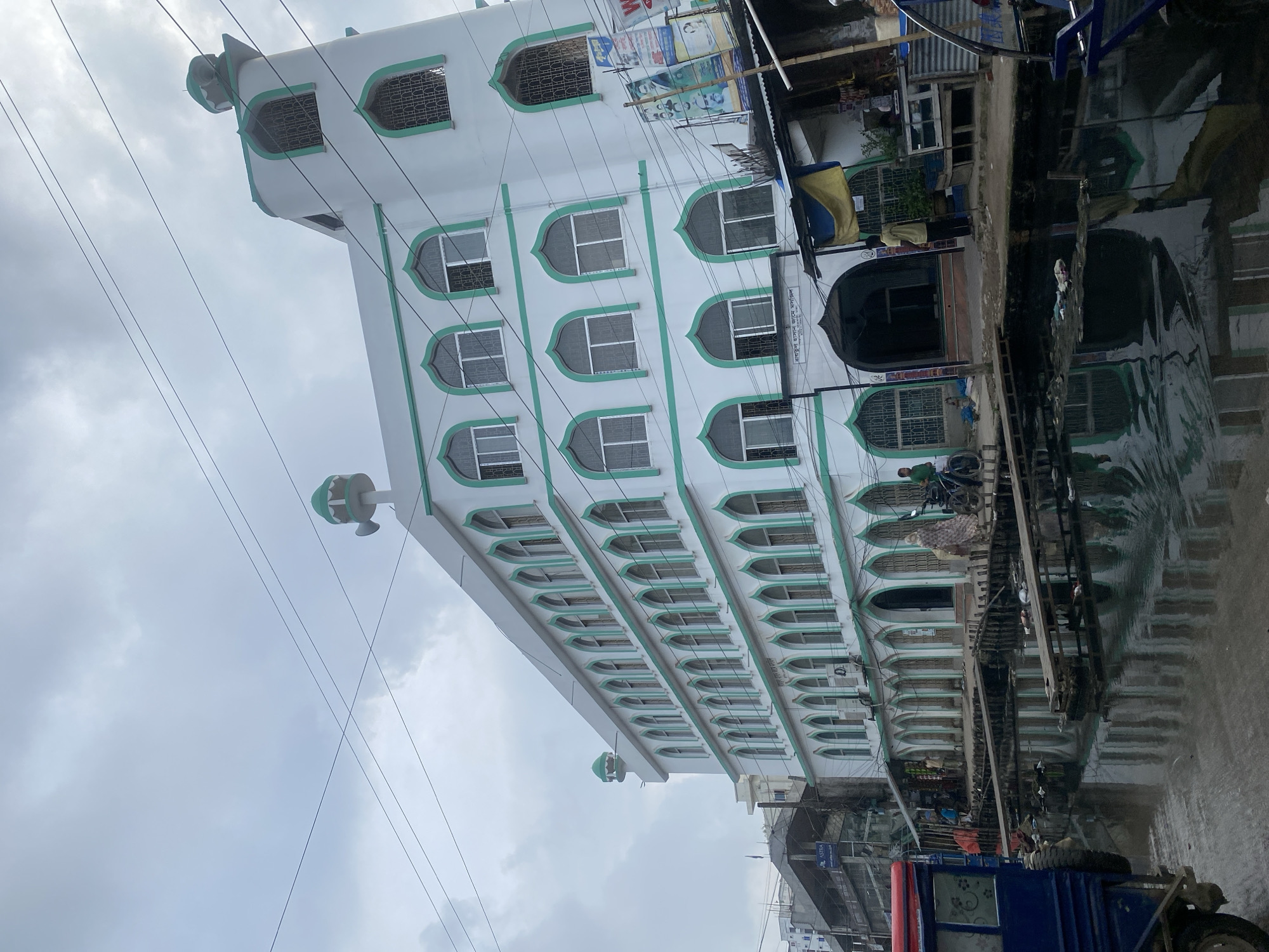 The image shows a multi-story white building with green trim, identified as the "Barakullah Hasan Jame Masjid" by a sign above its entrance. The building has numerous arched windows with decorative green outlines. The mosque is situated in Dhaka, Bangladesh, as indicated by the signage and architectural style, and appears to be located in a densely populated urban area. In front of the mosque, a murky canal or waterway reflects the building and the overcast sky. The canal is filled with debris, and makeshift wooden walkways are constructed over it. To the left of the canal, a blue vehicle, possibly a truck or cart, is partially visible. Several people are present: a woman in a patterned dress and headscarf walks away from the camera, and a man in a green shirt on a bicycle is to her left. Another man in a yellow shirt stands near the entrance of the mosque. The scene suggests a busy, somewhat weathered urban environment typical of parts of Dhaka, with signs of infrastructure challenges like the canal. The sky is overcast, contributing to a diffused light.
