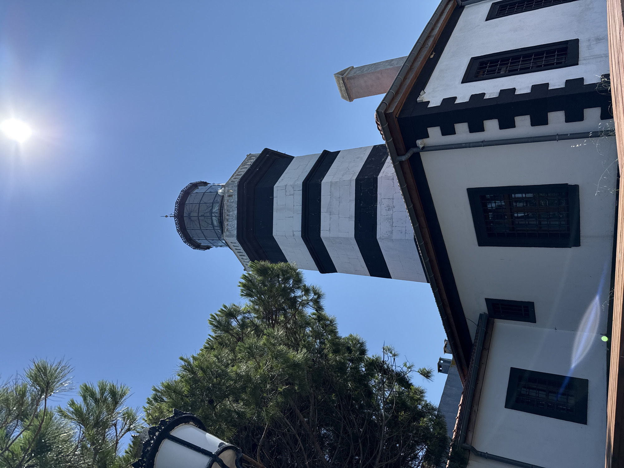 The image shows a lighthouse in Şile, Turkey, under a bright blue sky with the sun visible and creating a lens flare. The lighthouse is tall and octagonal, with alternating black and white bands. It is topped with a glass lantern room. Adjacent to the lighthouse is a white building with black trim and dark-paned windows. A chimney rises from the roof of the white building. In the foreground to the left, a portion of a decorative lamppost and some green pine branches are visible. The scene is viewed from a low angle, looking up towards the lighthouse and the sky.