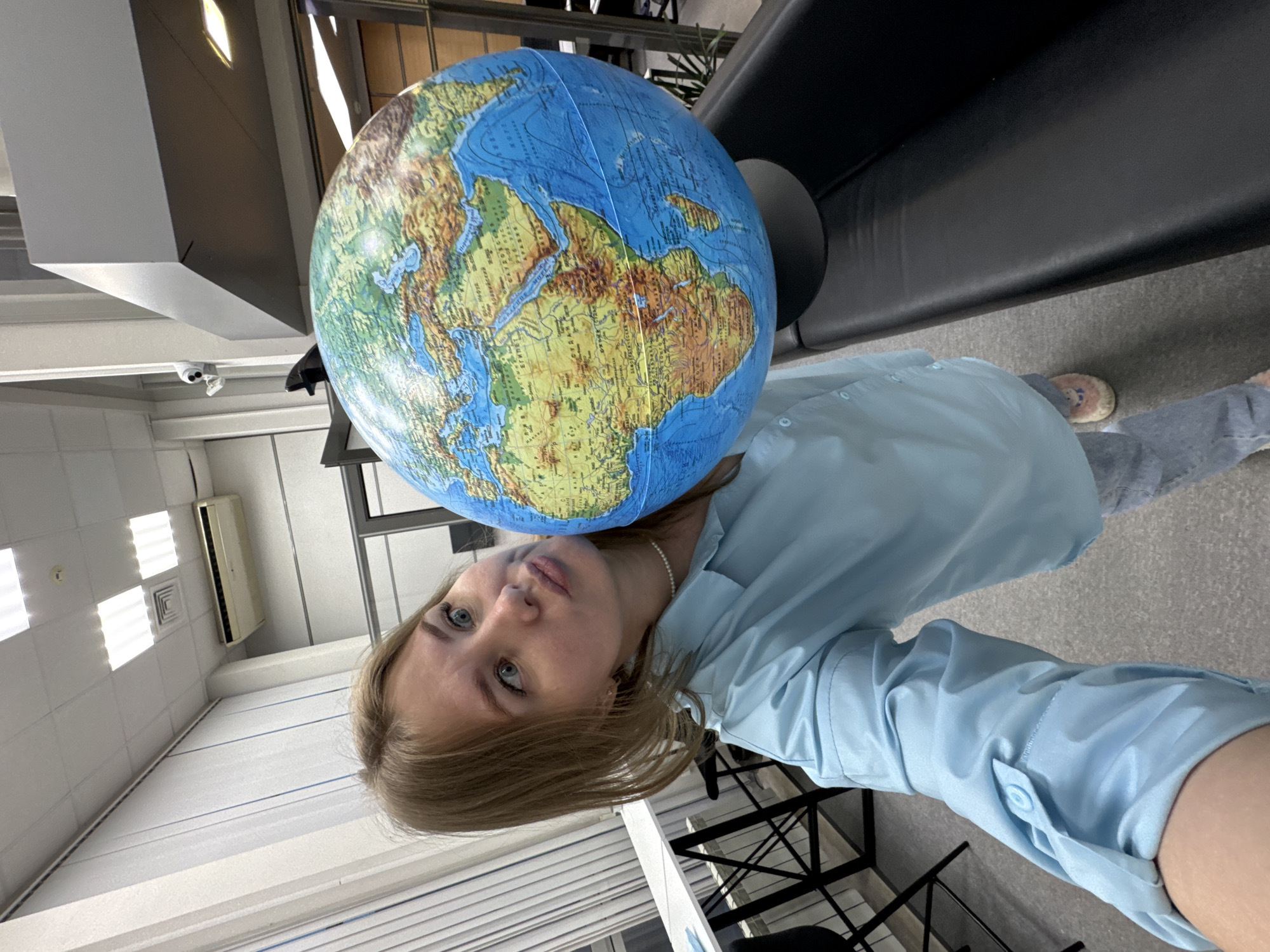 A woman in a light blue shirt stands indoors, holding a globe on her shoulder. The globe displays a map of Africa and Europe. She wears a pearl necklace. The location appears to be an office or institution with white walls, blinds on the window, and an air conditioner. Furniture includes a black couch and metal chairs. A security camera is mounted on the ceiling. The city of Perm, Russia is not discernible in this image.