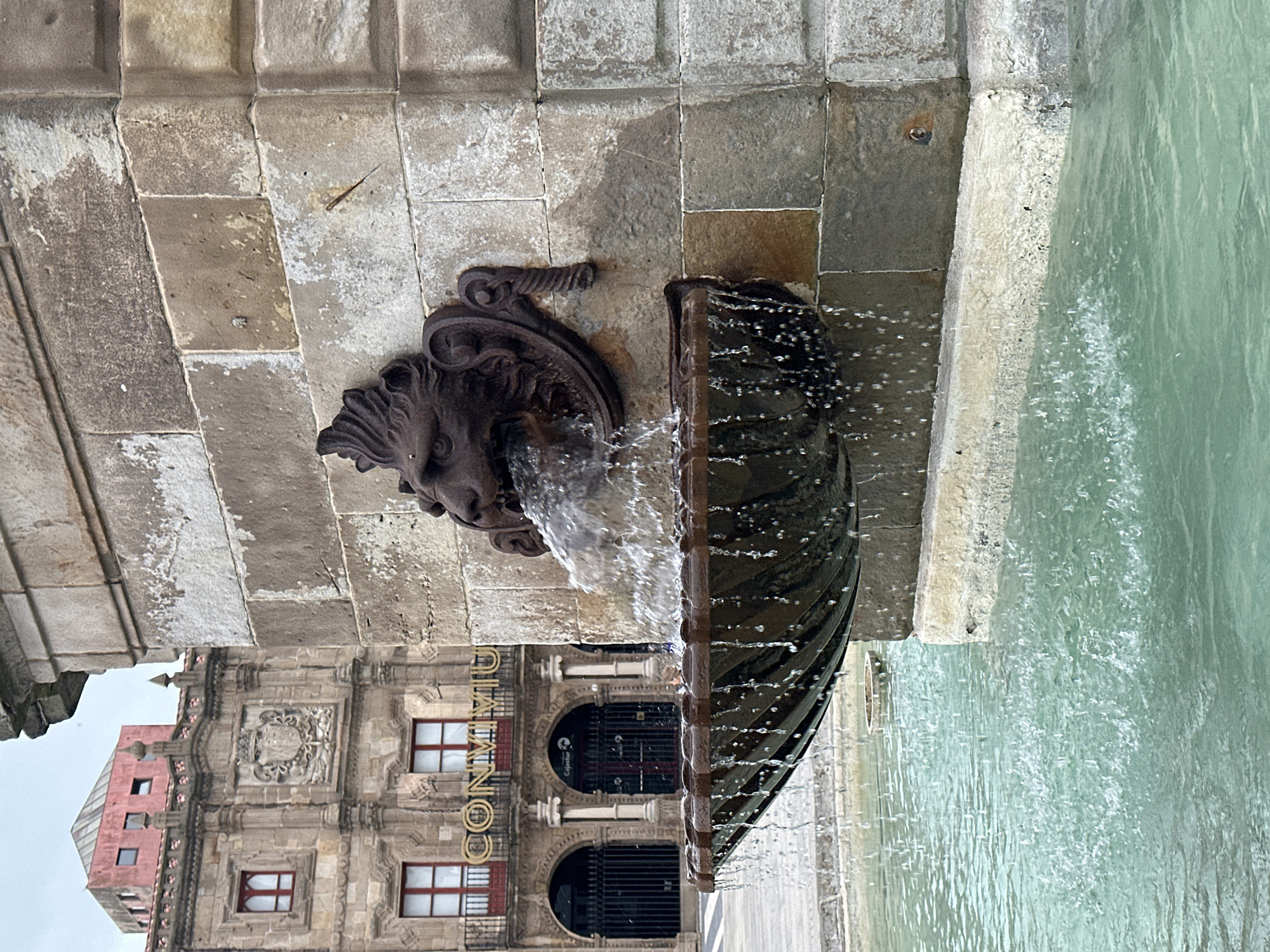 A lion-head fountain spouts water into a pool. The fountain is attached to a stone wall. Behind the fountain, a building with the name "CONMIMU" on it can be seen. This is located in the city of Gijón/Xixón, Spain.
