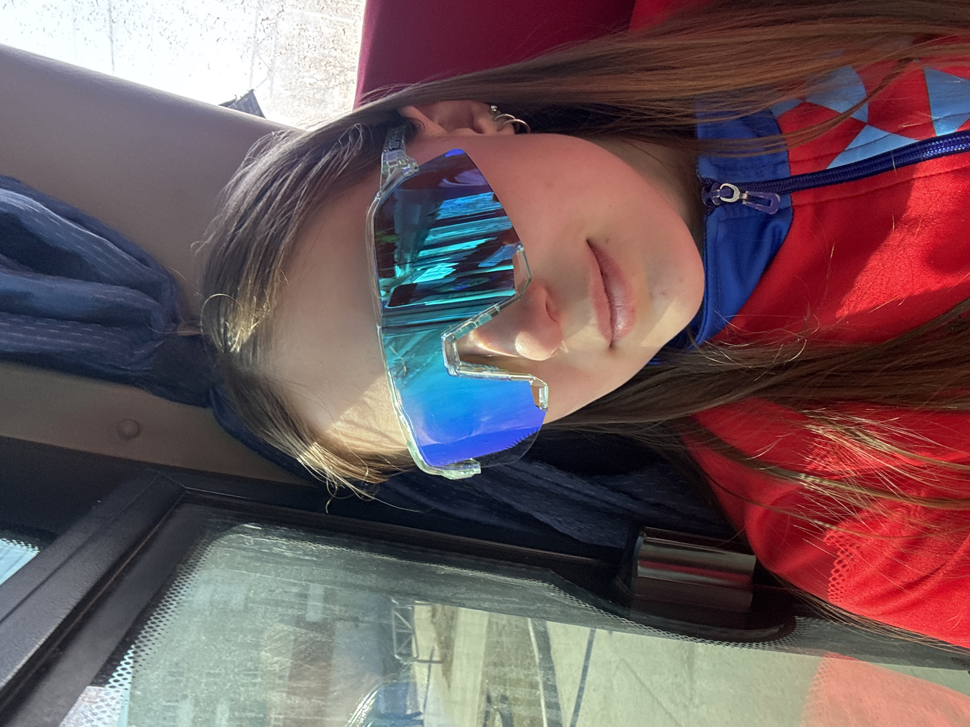 A woman wearing large, mirrored sunglasses and a red and blue jacket is in a vehicle. The windshield and dashboard of the vehicle are visible. The location is not definitively identifiable as Berezniki, Russia based on the image alone.
