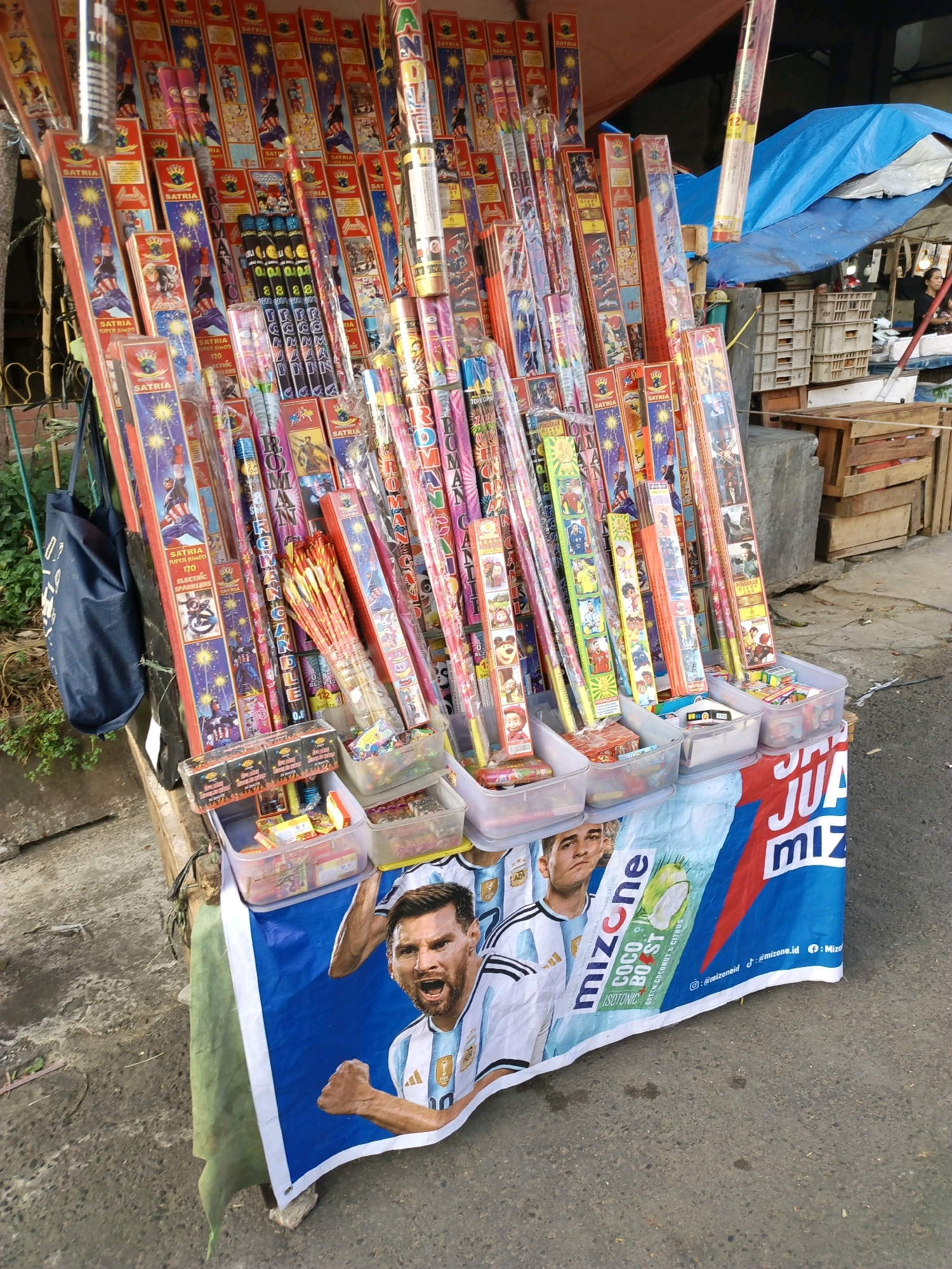 The image depicts a street vendor stall in South Tangerang, Indonesia, selling a variety of fireworks. The stall is covered by a red and blue tarp. The fireworks are arranged in vertical stacks, with many featuring colorful packaging depicting cartoon characters and superheroes. Some fireworks are bundled together in plastic sleeves, while others are loose. In the foreground, the stall is adorned with a banner featuring images of soccer players, specifically Lionel Messi, and the brand "Mizone". Several transparent plastic containers filled with smaller fireworks or firecrackers are placed on the lower shelves of the stall. The ground is paved with asphalt, and there are some plants and a blue bag visible on the left side of the image. In the background, there are stacks of wooden crates and a blue tarp covering part of the stall. A woman is partially visible in the background, standing near the crates.