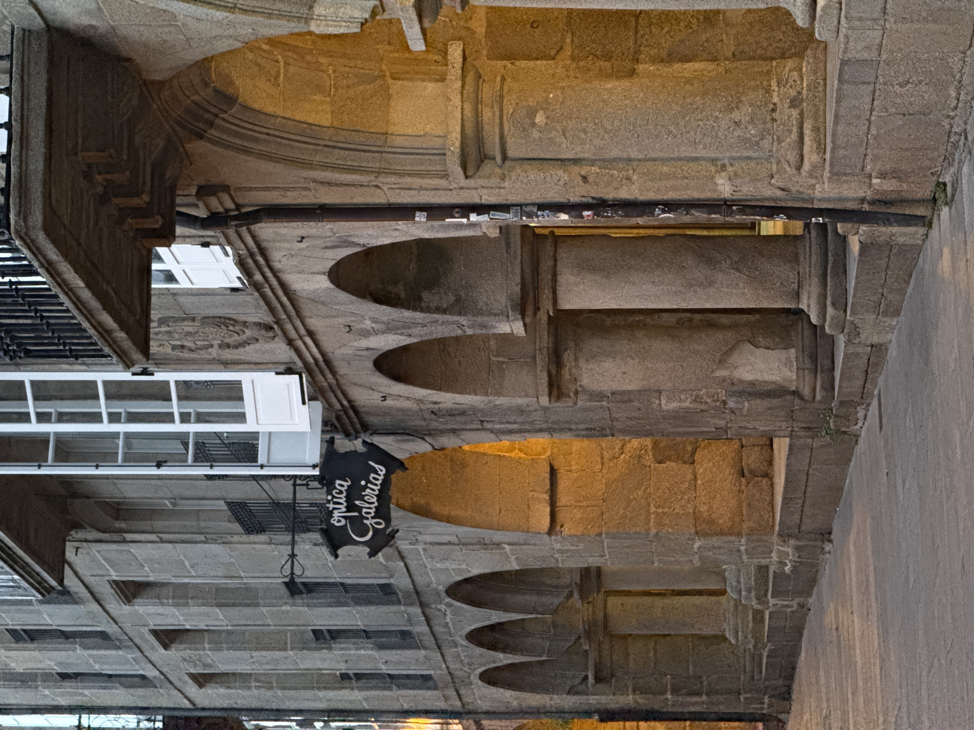 The image shows a view of the Rúa do Franco in Santiago de Compostela, Spain. It is an outdoor scene with a focus on the architecture of the buildings lining the street. The buildings are constructed from stone, with arched doorways and balconies. The light suggests it is either early morning or late evening, with a warm glow emanating from within some of the arched openings. A sign with the words "optica galerias" is visible hanging from one of the buildings. The street itself appears to be paved with stone. There are no people visible in the image. The overall impression is one of historical charm and a quiet urban streetscape.