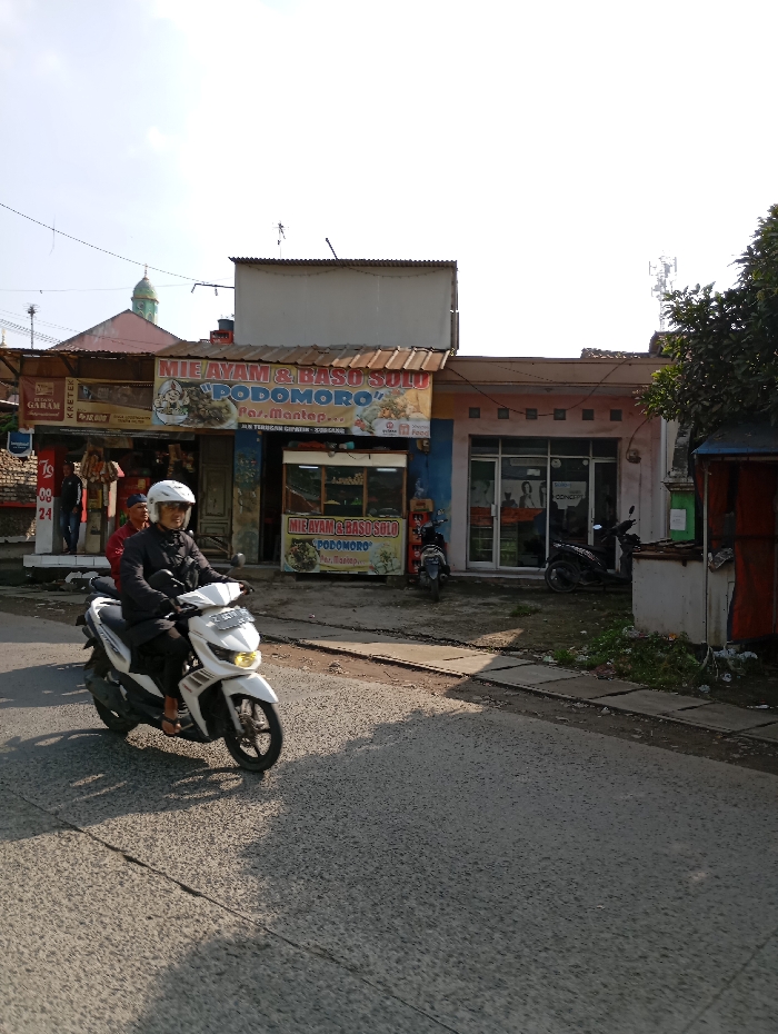 The image captures a daytime street scene in Pataruman, Indonesia. An asphalt road occupies the foreground and midground, exhibiting textural variations and shadows indicative of overhead sunlight.
A white Yamaha Mio scooter, bearing license plate 'Z 3670 QO', is prominently positioned in the left-center, captured in motion from left to right. The rider, wearing a white full-face helmet and a dark jacket, is operating the vehicle.
To the right of the road, a concrete sidewalk abuts a row of commercial buildings. The central building is a two-story structure with a brown sloped roof extending over its ground floor. A large yellow banner affixed to its facade advertises "MIE AYAM & BASO SOLO PODOMORO," displaying text and food imagery. Below this, a glass display counter is visible. Two individuals are standing near the entrance of this establishment; one is partially obscured, wearing a red shirt. To the immediate left of this shop, a narrower storefront is partially visible, featuring a banner for "GARAM KRETEK" with "Rp 18.000" displayed.
Adjacent to the "Podomoro" shop on its right, another single-story building with a light-colored, possibly pink or peach, facade features multiple glass windows or doors. Two scooters, one black and one dark grey, are parked on the sidewalk in front of this building. A tree with green foliage partially frames the right edge of the scene.
In the background, a green-domed structure, characteristic of a mosque, is visible above and behind the leftmost building. Overhead utility lines traverse the clear sky. Further right in the background, a cellular tower or telecommunications mast is discernible against the bright horizon.