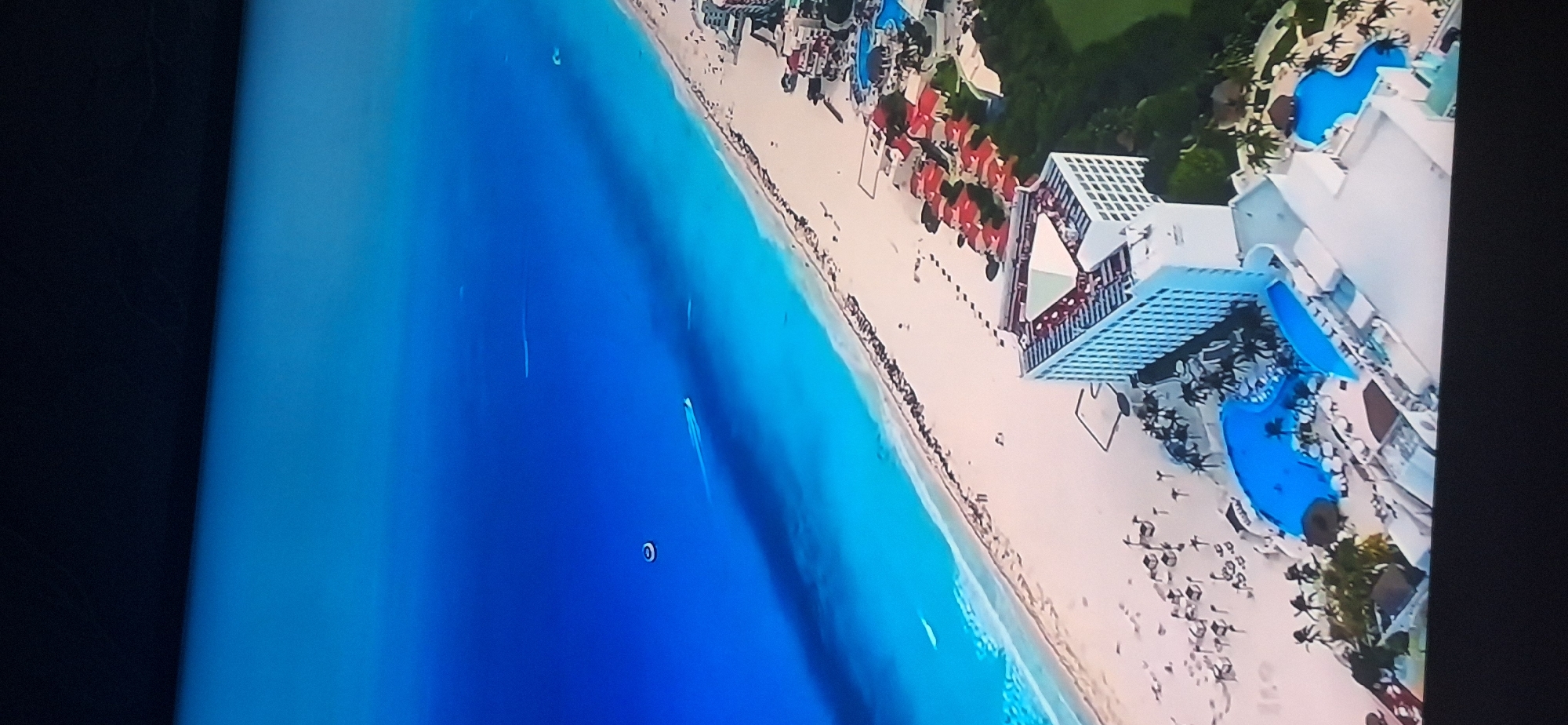 This is an aerial view of a coastal resort. The ocean is a vibrant blue, transitioning to turquoise near the shore where it meets a sandy beach. There are several boats leaving white wakes in the water. The beach is dotted with beach chairs and umbrellas. A large, modern hotel complex with a triangular roof structure is visible, along with several smaller buildings with red roofs. There are swimming pools, some filled with blue water, and palm trees are scattered throughout the resort. This scene is not located in Bila Tserkva, Ukraine, as Bila Tserkva is an inland city.
