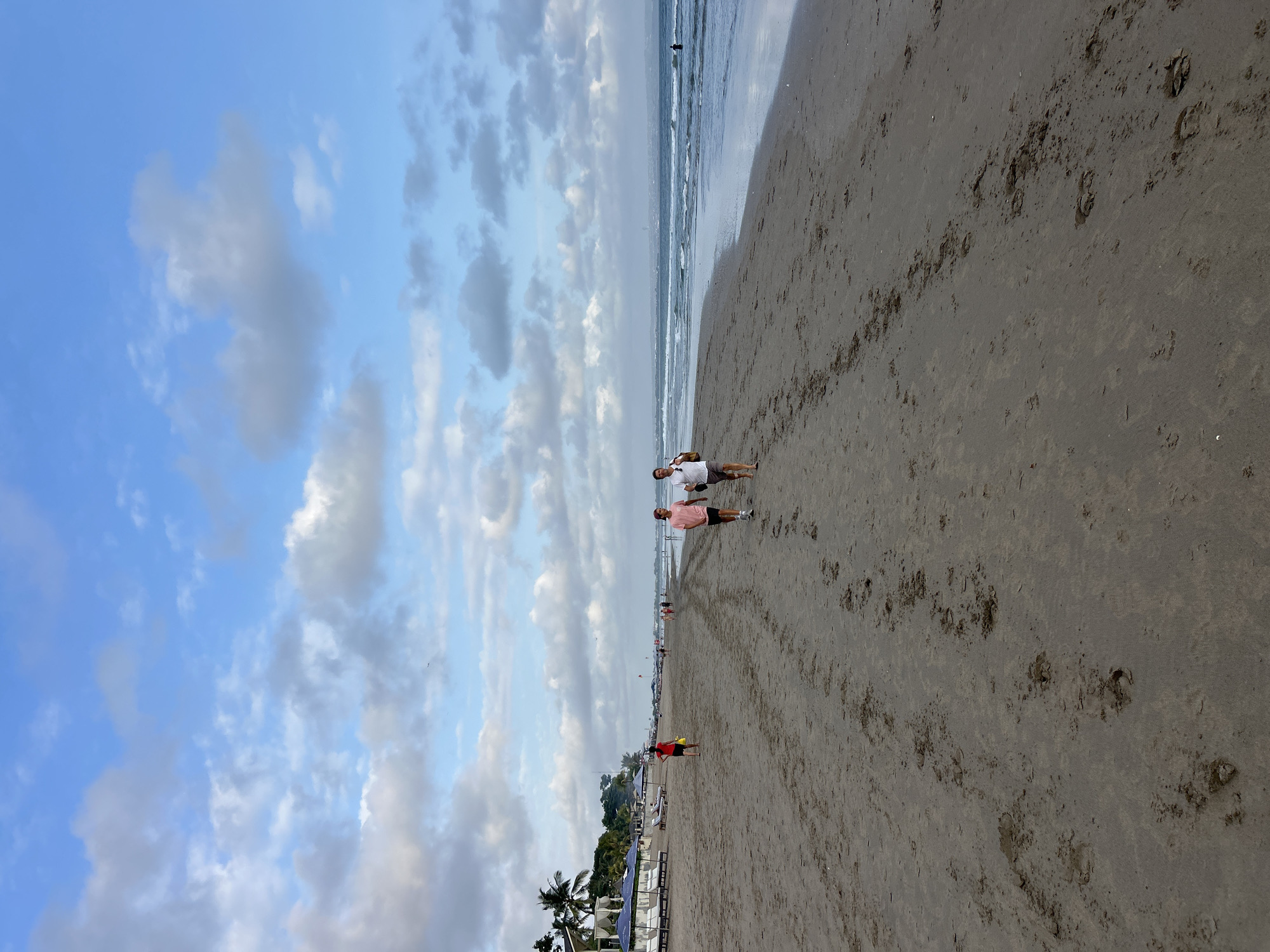 A wide, sandy beach is depicted under a sky with scattered clouds. In the foreground, numerous footprints mark the wet sand. Two men are walking along the shore, one wearing a pink t-shirt and dark shorts, the other a white t-shirt and light shorts. Further down the beach, other people are visible walking, and a few beach chairs and umbrellas are set up on the left side. Palm trees and buildings line the edge of the beach in the distance. The ocean stretches out to the horizon, with gentle waves breaking on the shore. The scene appears to be located in North Kuta, Indonesia.