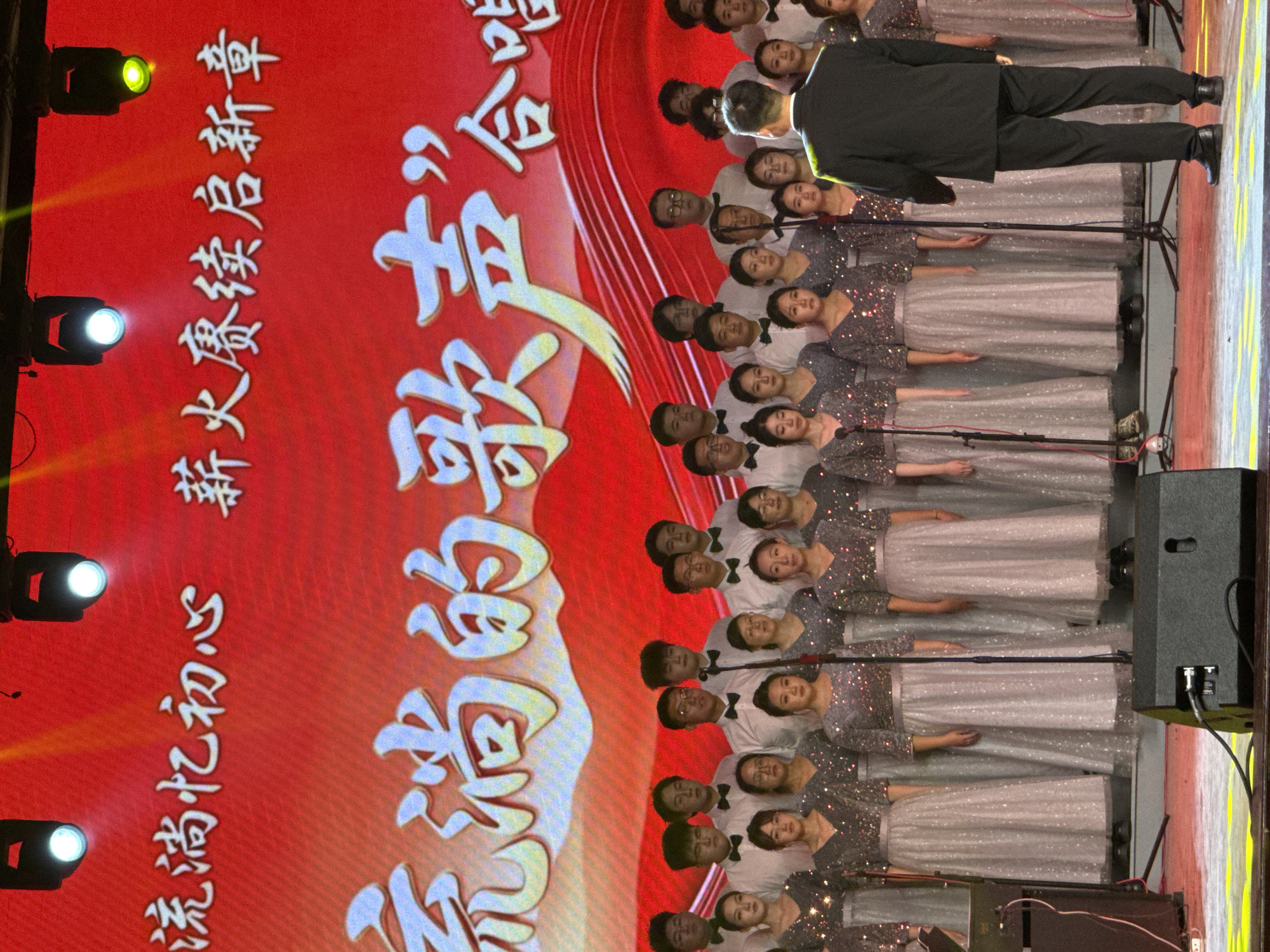 This image captures an indoor choir performance or rehearsal, likely taking place in Guta, China. The scene is dominated by a large red backdrop or screen displaying prominent white Chinese characters. The main text, in a large, stylized font, reads "流淌的歌声”合唱" which translates to "'Flowing Songs' Choir" or "Chorus." Above this, another line of text says "流淌忆初心 薪火赓续启新章," which can be interpreted as "Flowing, remembering the original aspiration; the torch is passed, starting a new chapter."
A large choir, comprising both men and women, is arranged in multiple rows, facing away from the viewer towards the backdrop. The women are dressed in elegant, light-colored, possibly silver or sequined gowns with long skirts, while the men wear white shirts, black bow ties, and black trousers. Many choir members have individual microphones or shared microphones positioned in front of them. In the foreground, with his back to the viewer, stands a conductor dressed in a dark suit, actively leading the choir.
The stage is illuminated by several spotlights, some casting bright white light and others a yellow-green hue, suggesting a performance setting. To the right of the choir, stage equipment such as large black speakers and cables are visible. The expressions of the choir members appear focused and engaged in the singing. The overall atmosphere suggests a formal and well-prepared musical event.