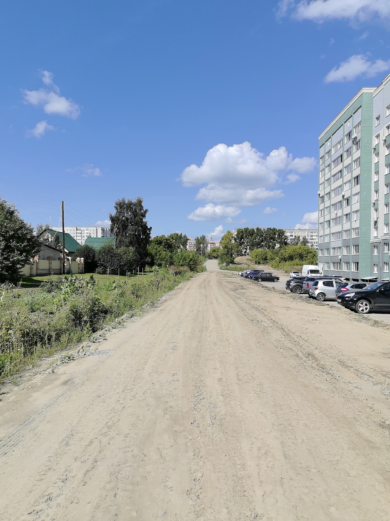 The image shows a dirt road leading away from the viewer, with a modern apartment building on the right and a residential area with smaller houses and trees on the left. The sky is blue with scattered white clouds. Several cars are parked along the right side of the road, near the apartment building. In the distance, more buildings and trees are visible. The overall impression is a suburban or semi-urban environment under a clear sky. This scene is consistent with areas in Novoaltaysk, Russia, which features a mix of modern apartment complexes and older, smaller residential areas alongside unpaved roads.