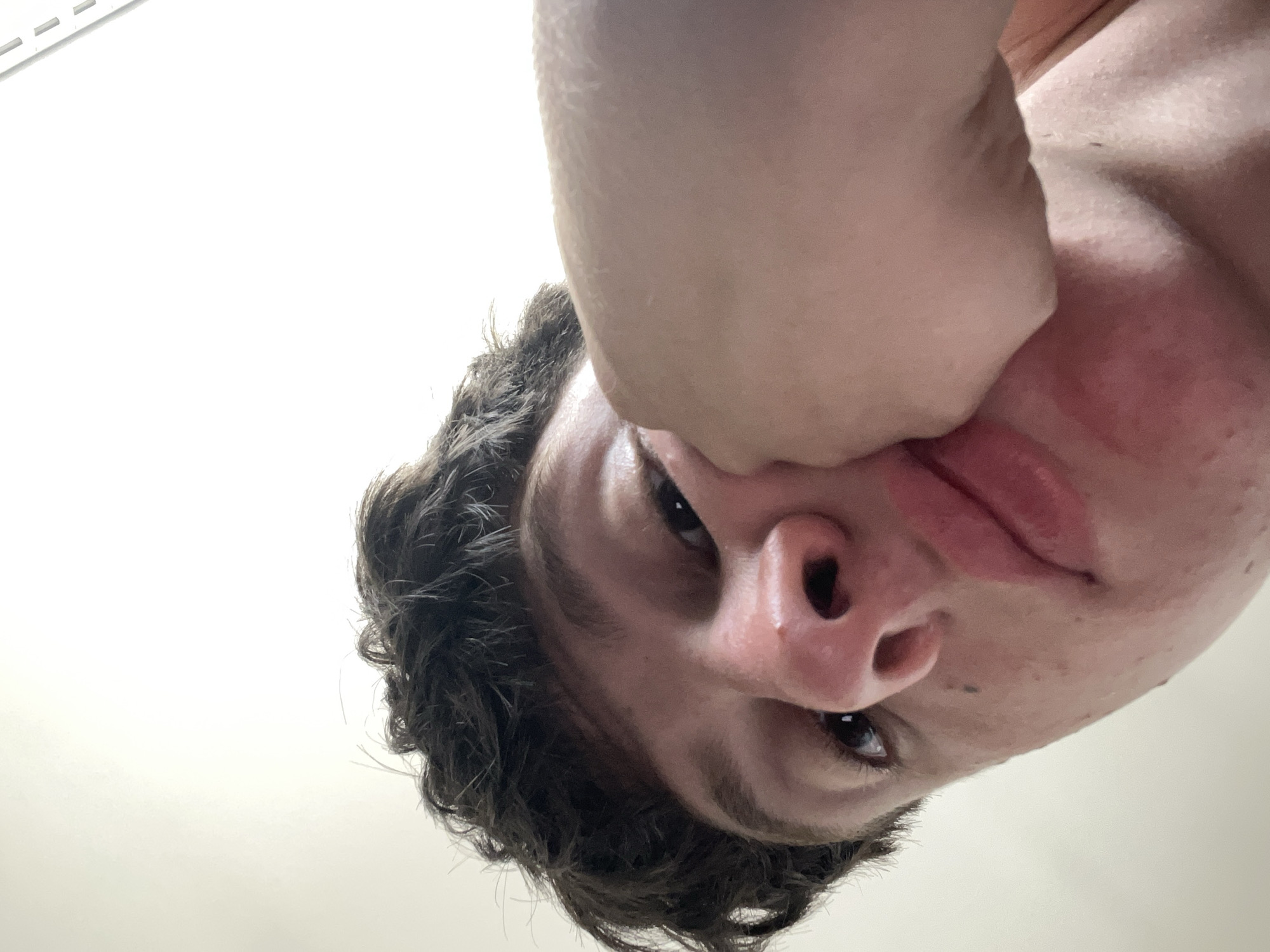 A close-up, low-angle shot shows a young person with dark, curly hair resting their cheek on their hand. Their eyes are open, and their mouth is slightly agape, revealing a glimpse of their teeth. The lighting is bright, coming from above, casting minimal shadows. The background is a plain, light-colored wall, possibly a ceiling or a blank wall. There are no discernible details about the environment that would indicate a specific location within a city. The image appears to be a selfie, taken from an unusual perspective.