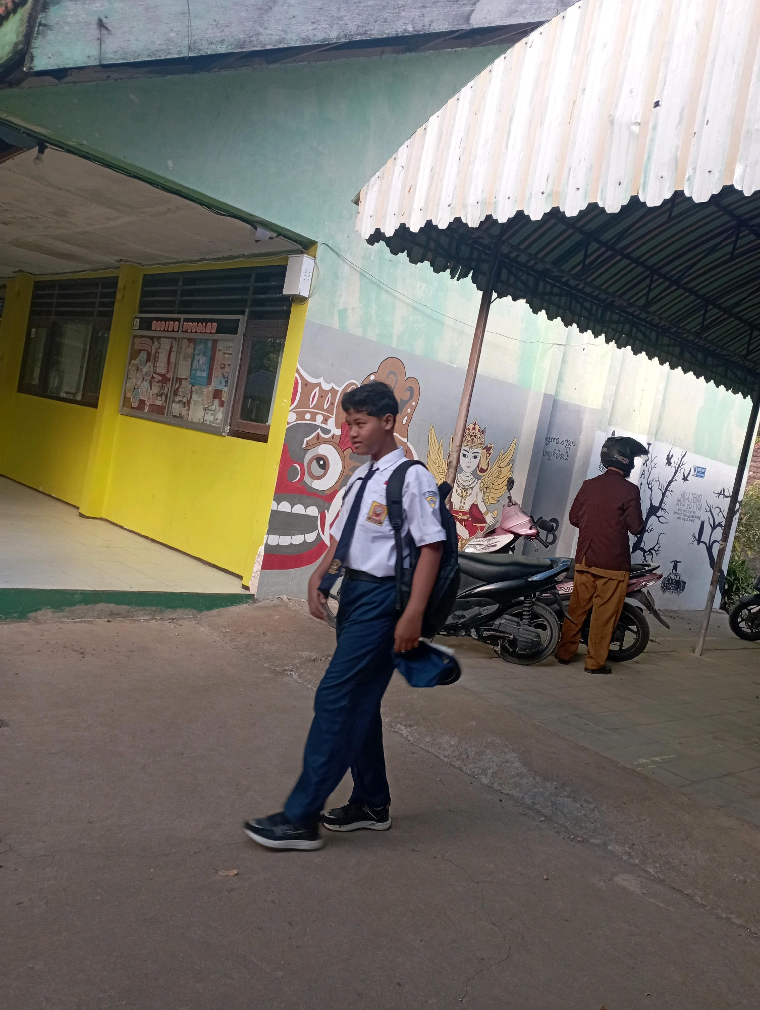 A young Indonesian male student in a white shirt and blue pants with a backpack walks on a paved path outside a yellow building with a green-painted upper wall. The student is looking to the right. To the right of the student, two motorcycles are parked. A person wearing a brown jacket and light brown pants with a black helmet is standing next to the motorcycles, facing away from the camera. Behind the motorcycles is a wall with colorful murals, including a depiction of a deity with wings and a large face with red and white patterns. The environment appears to be an outdoor area of a school in Banyuurip, Indonesia, indicated by the building's architecture, the school uniform, and the murals. The lighting suggests it is daytime.
